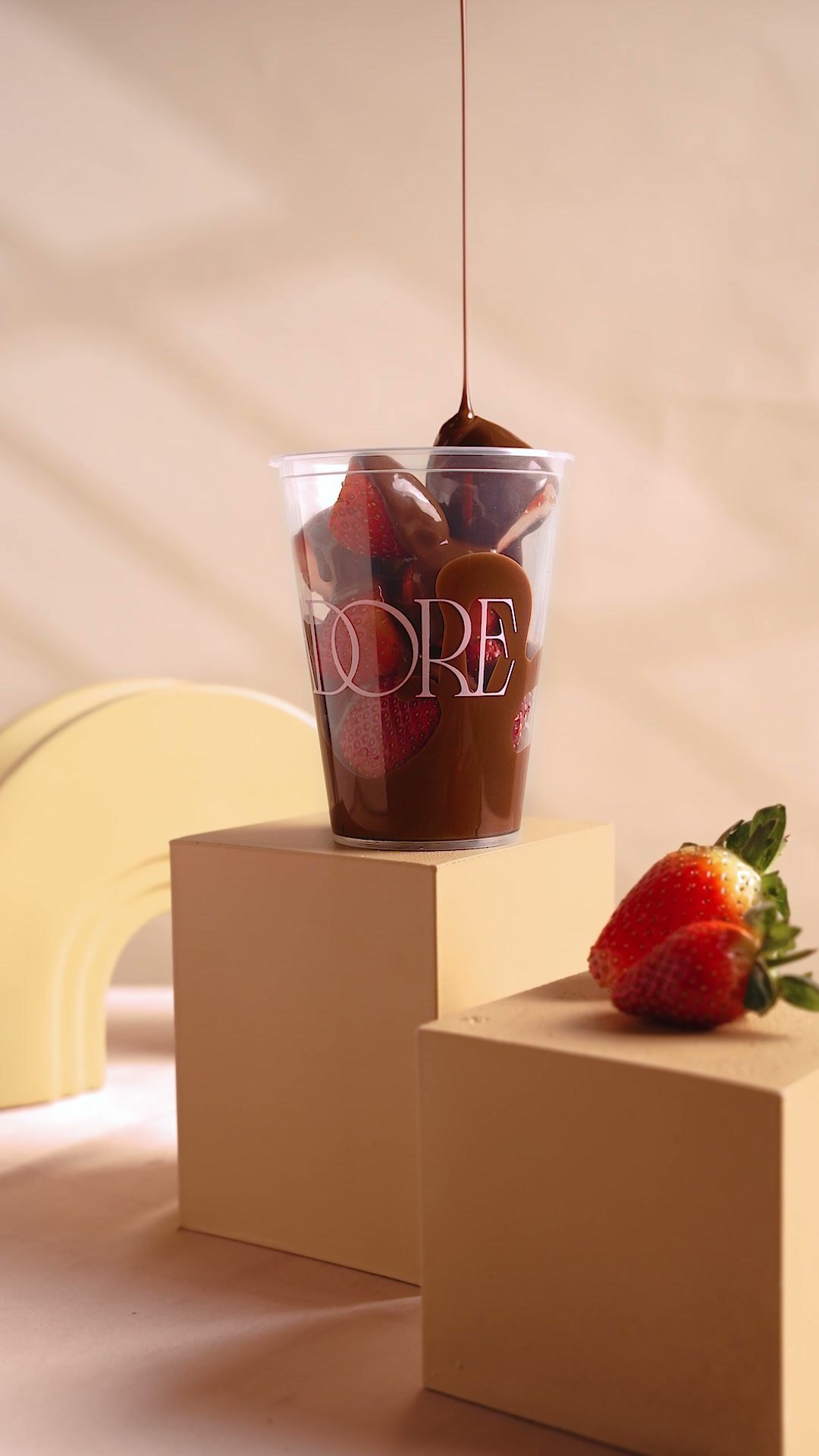 Chocolate sauce pouring into a cup of Adore chocolates on stacked plinths, a strawberry beside it