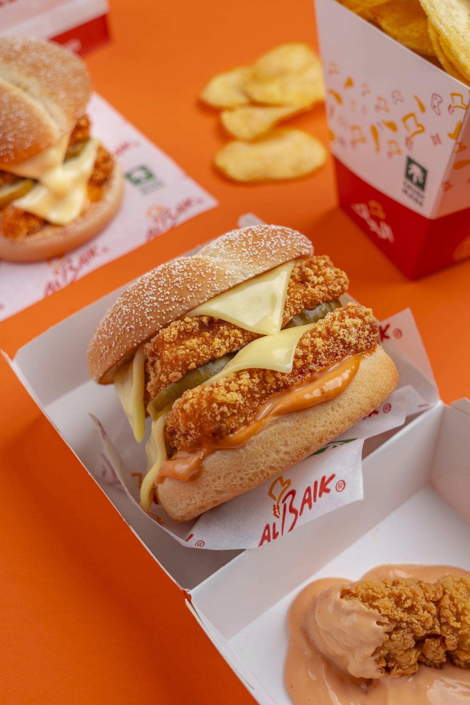 Crispy chicken and cheese burger open in its Al Baik box, fries and crisps behind it