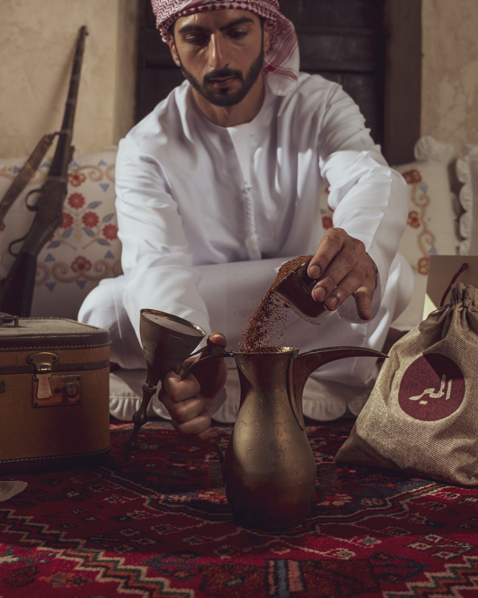 Man in a kandura pouring coffee from a brass dallah on a majlis carpet