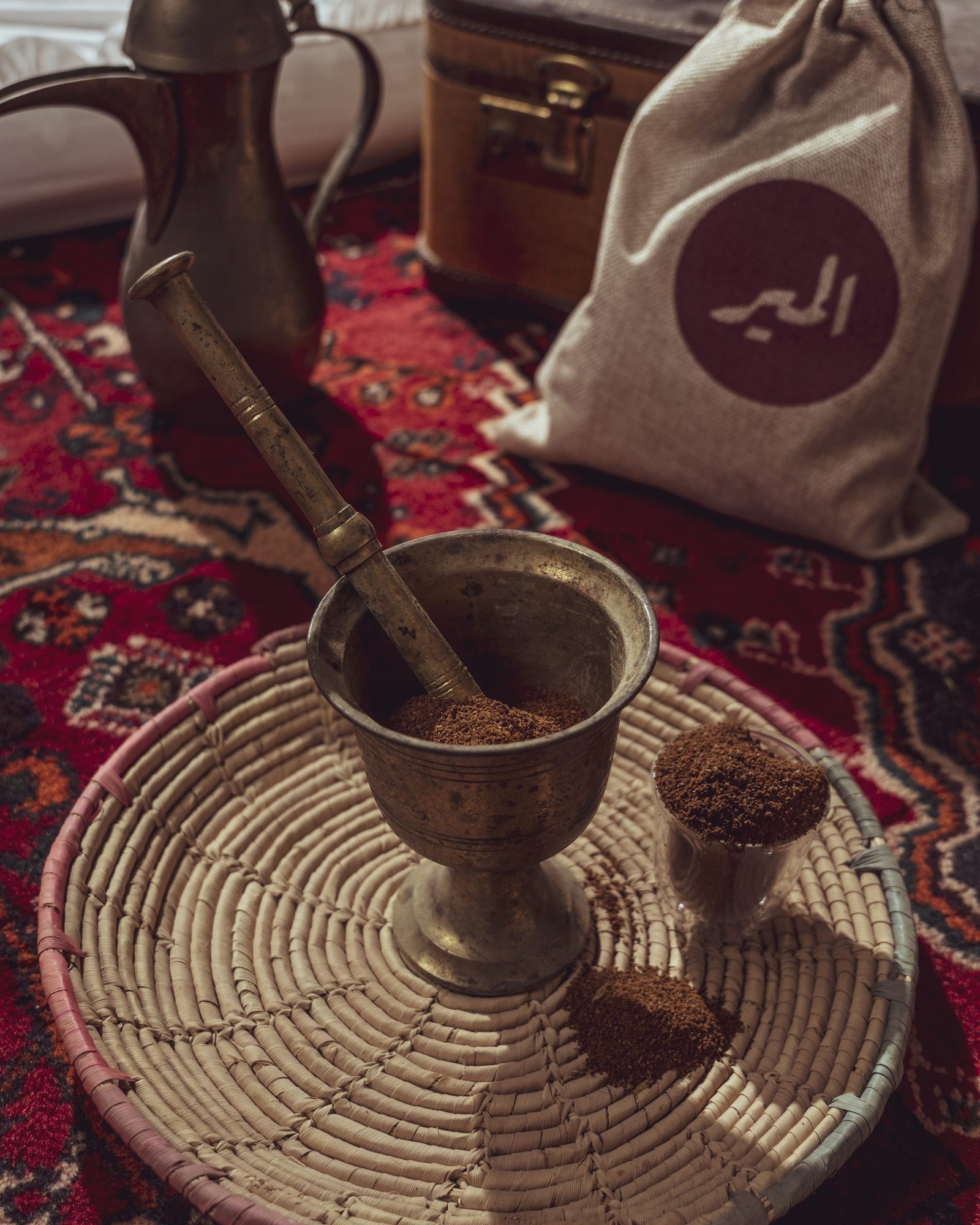 Brass mortar and pestle with ground coffee on a woven tray beside stamped sacks