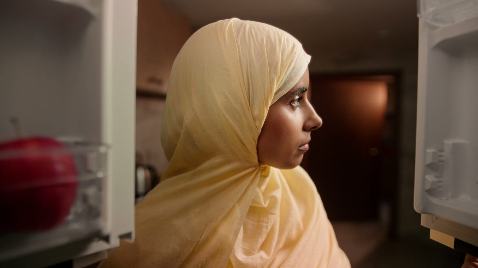 Woman in a yellow hijab in profile, standing at an open fridge