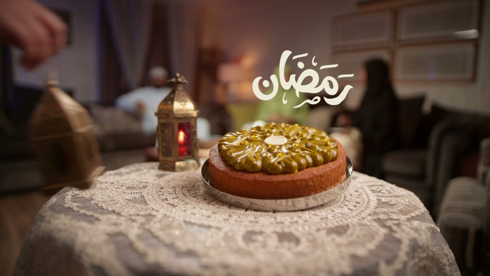 Pistachio-topped tart on a lace cloth beside a brass lantern, Ramadan lettering in the corner