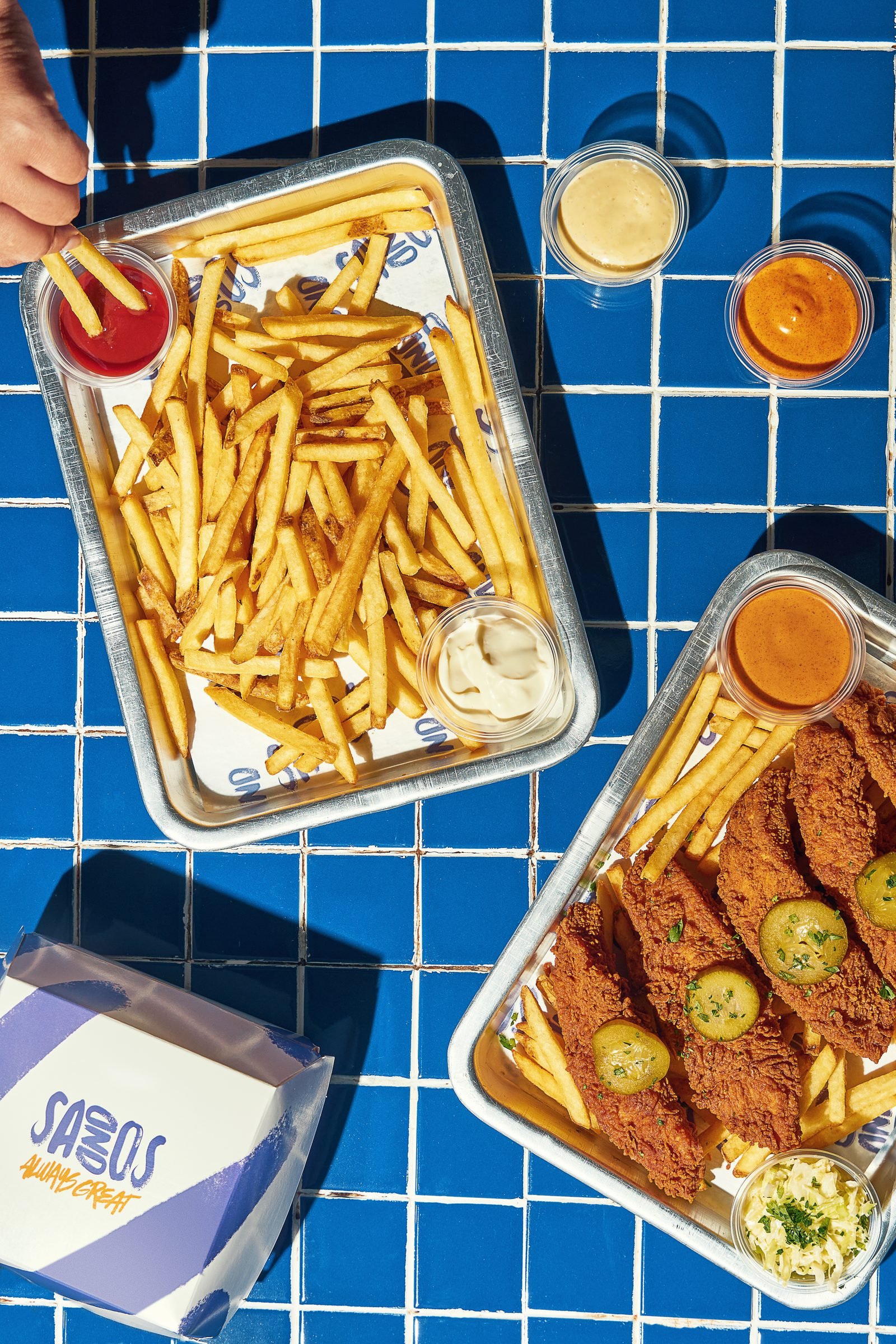 Two foil trays of fries and fried chicken with dips, on blue tiles