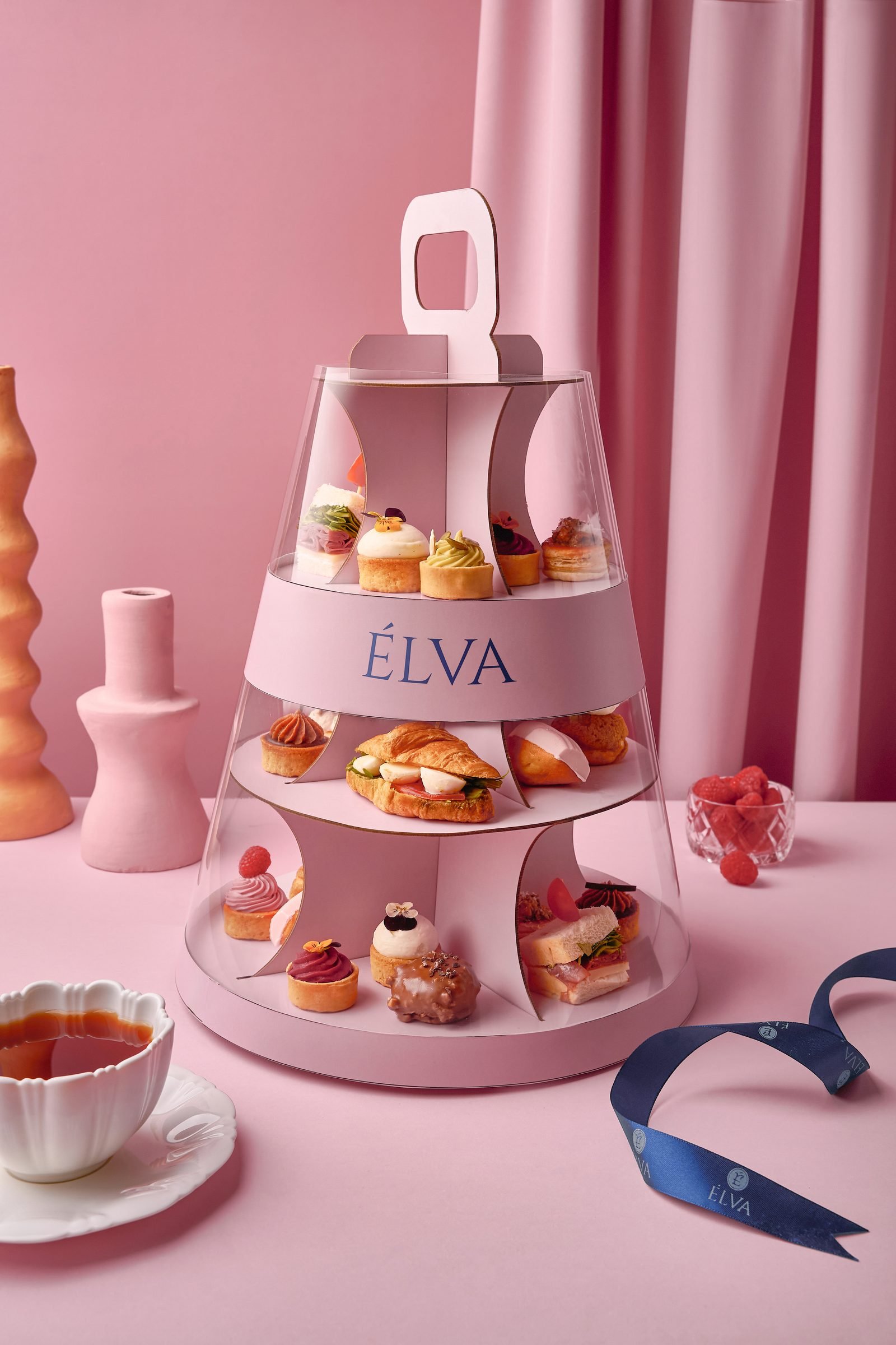 Three-tier Elva stand of petits fours and mini cakes with a cup of tea, on pink