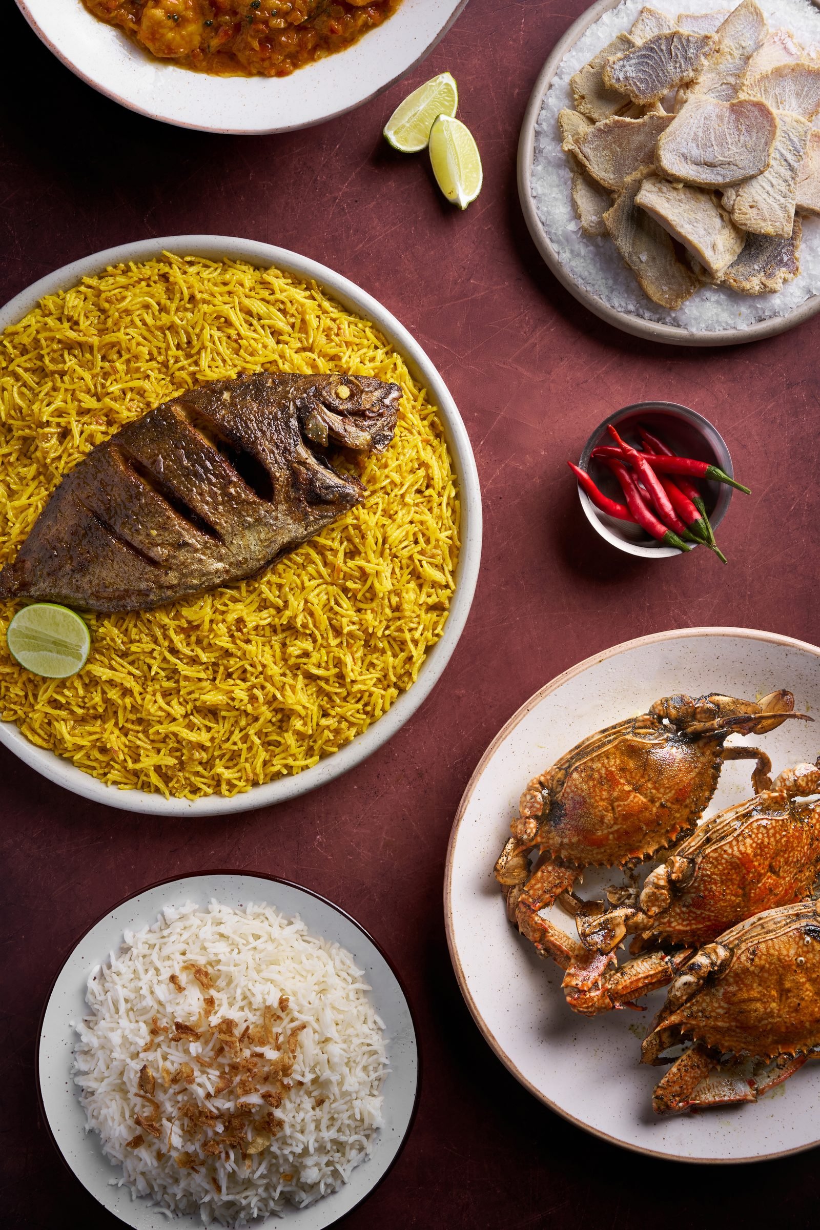 Overhead spread of whole fried fish on saffron rice, crab, white rice and grilled meat on deep red