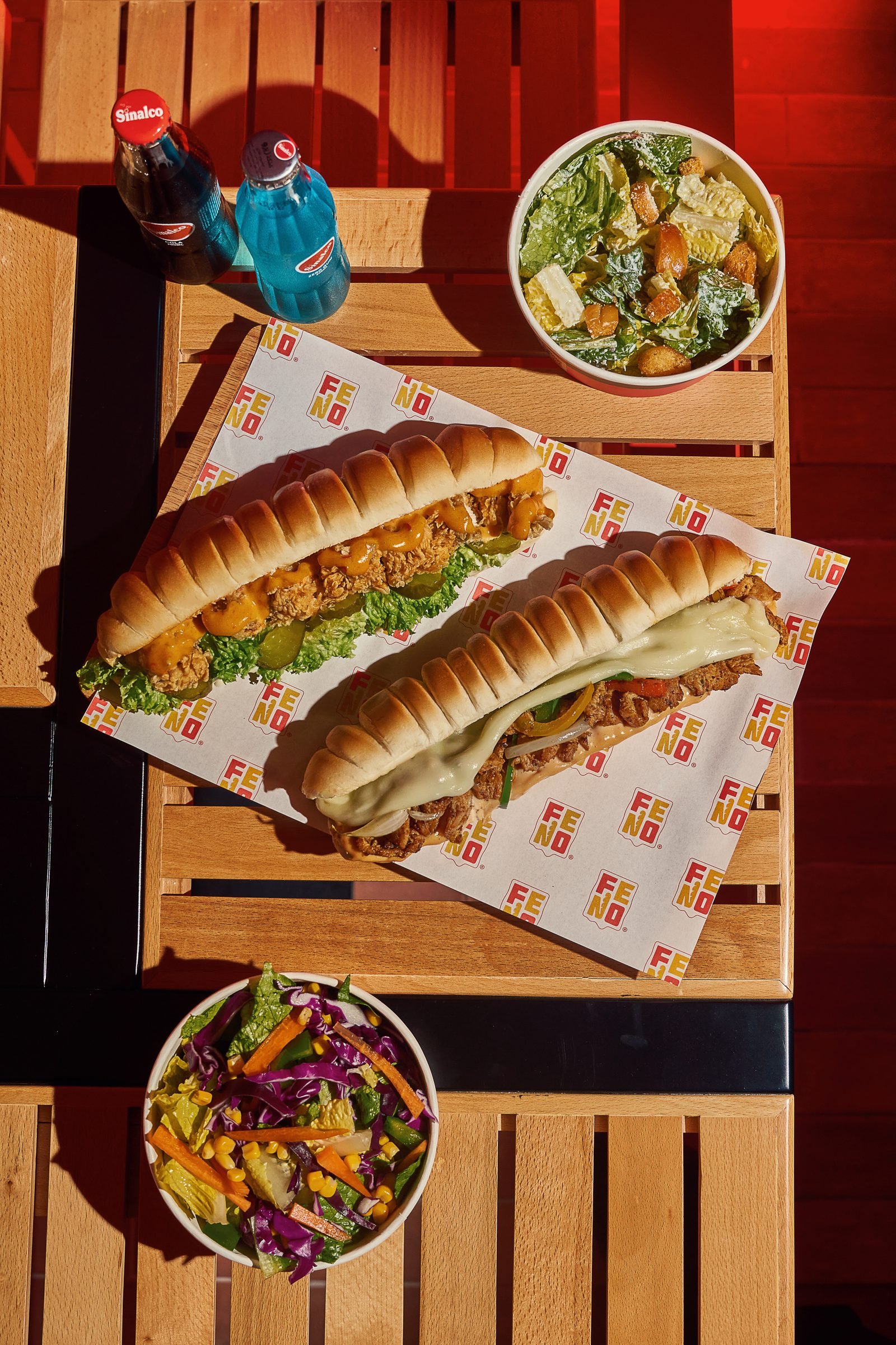 Two long submarine sandwiches on Fino paper on a wooden bench, with salads and a bottle
