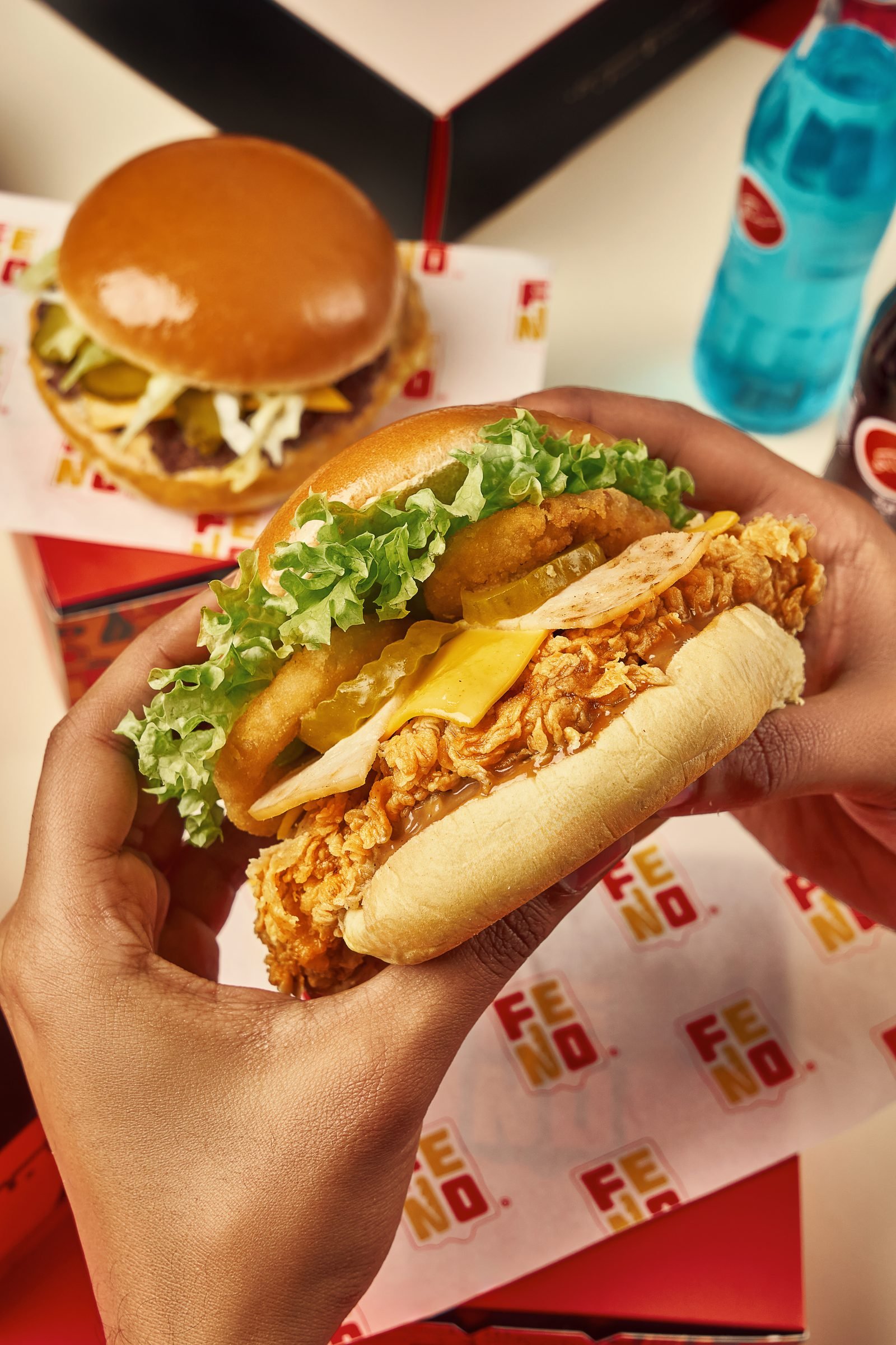 Hands holding a fried chicken sub over a table of Fino packaging and a bottle
