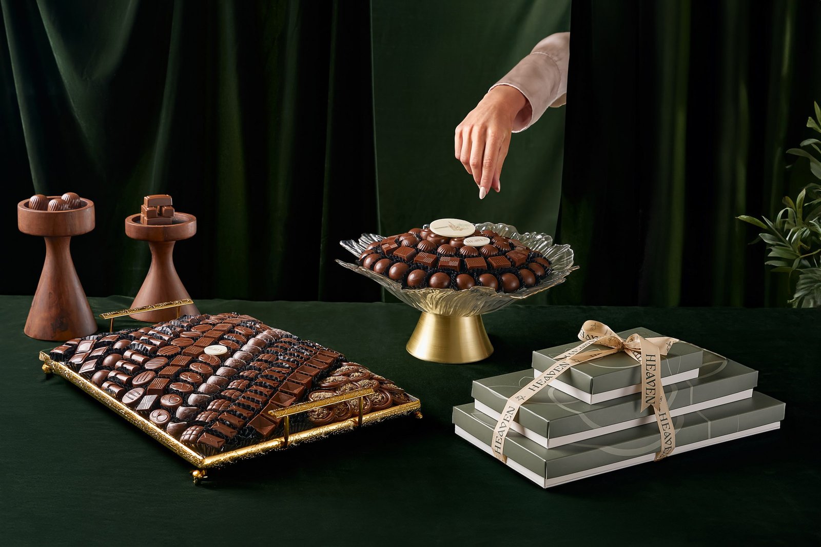 Dark green table of chocolate trays and gift boxes, a hand reaching for a truffle