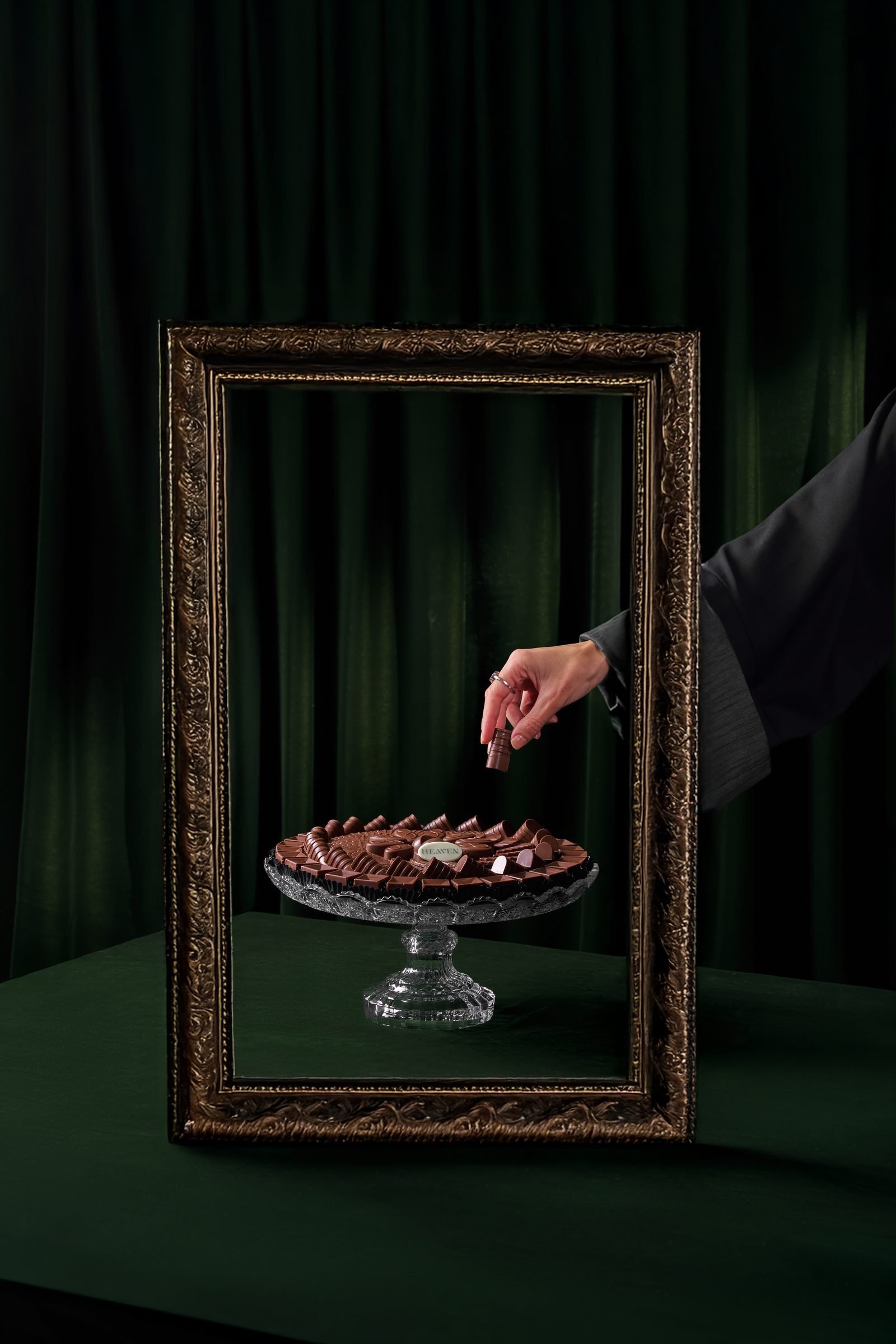 Hand taking a chocolate from a glass cake stand inside an empty gilt frame, on dark green