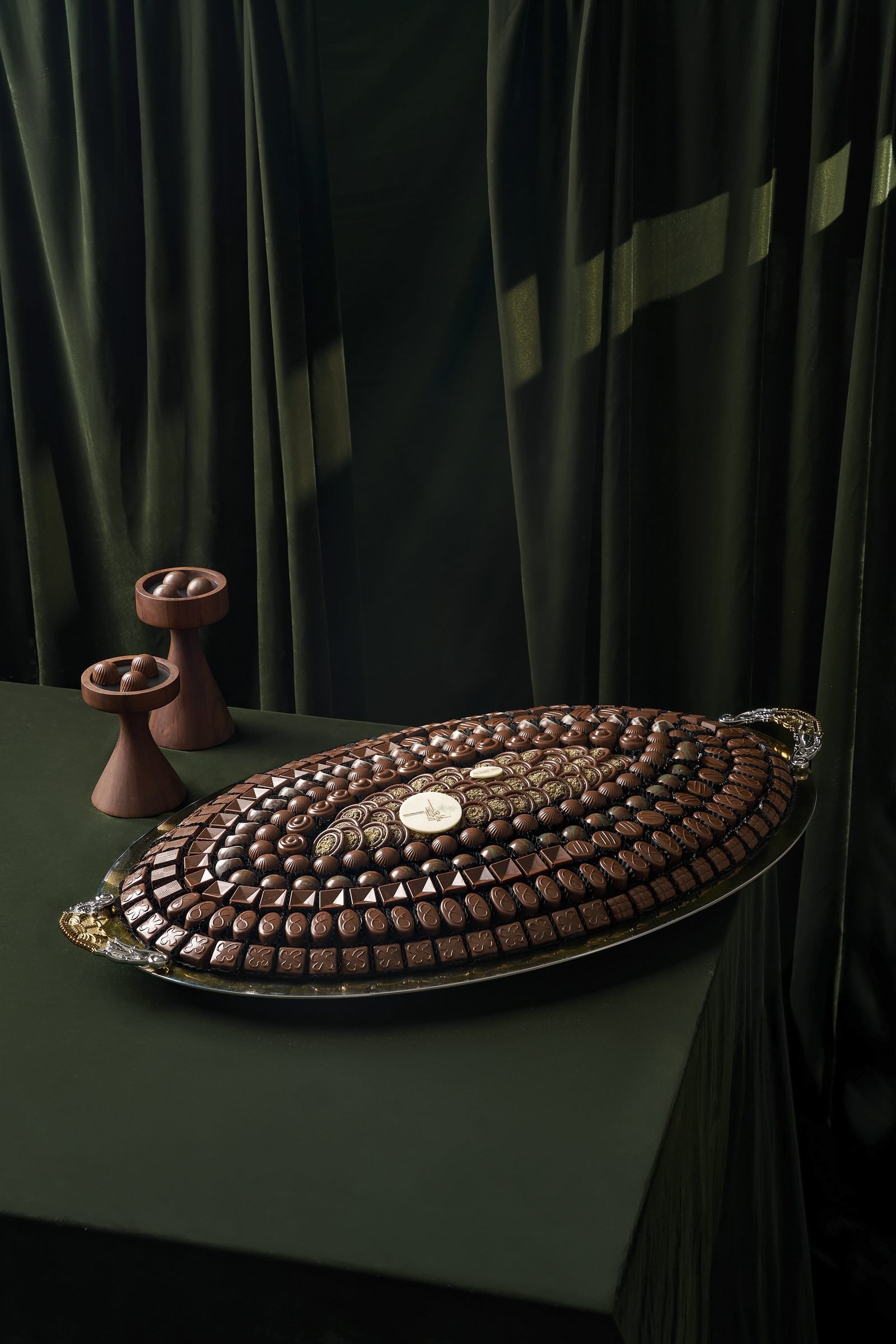 Oval platter of chocolate rows on a dark green table between two candle holders