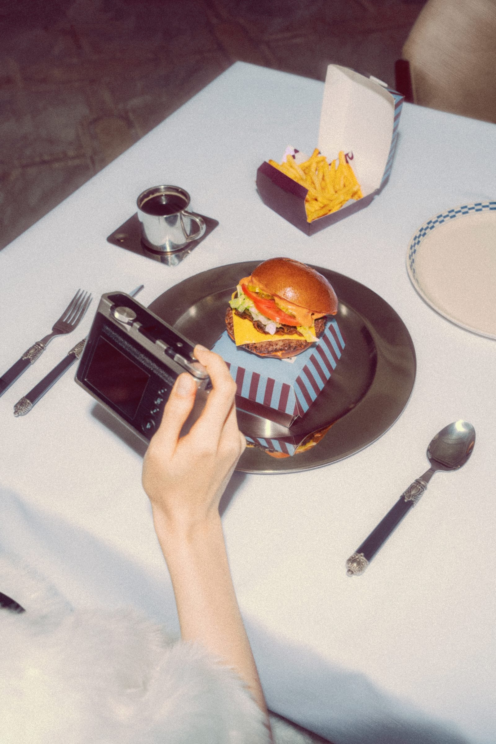 Hand reaching for a burger on a metal tray, a vintage camera beside it on a pale table