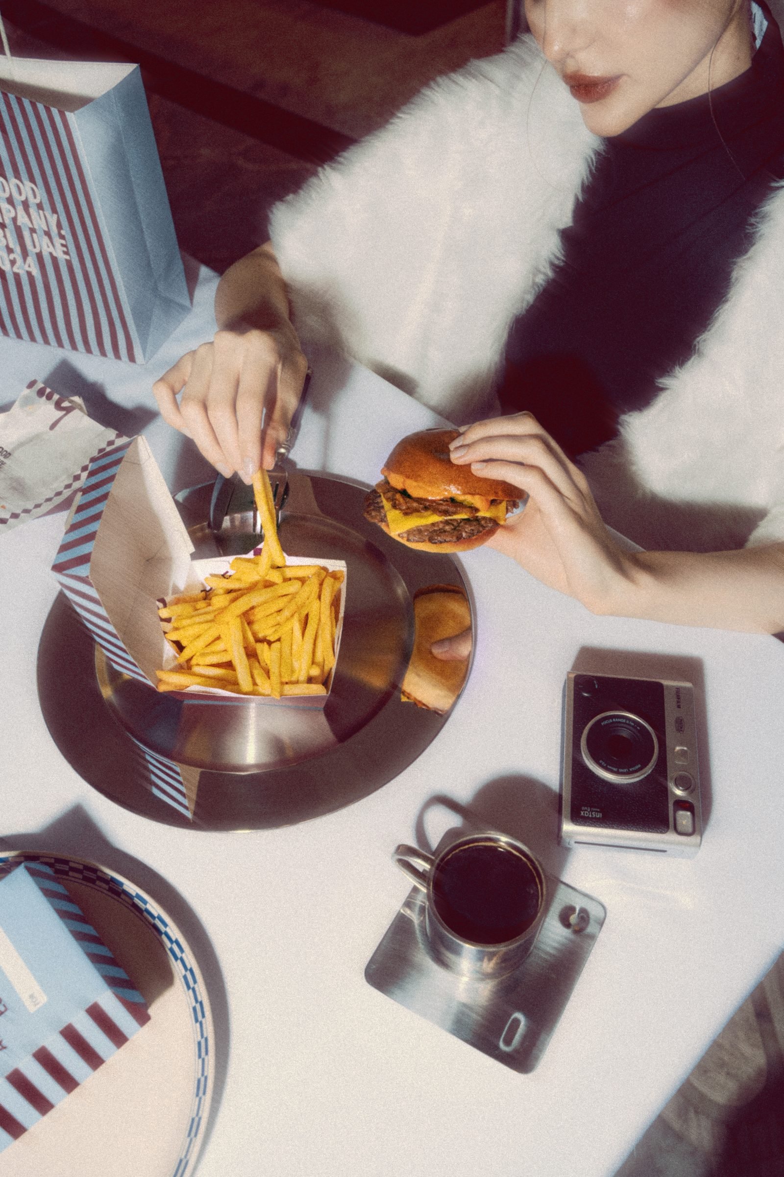 Woman in a fur coat eating a burger and fries at a set table with a film camera