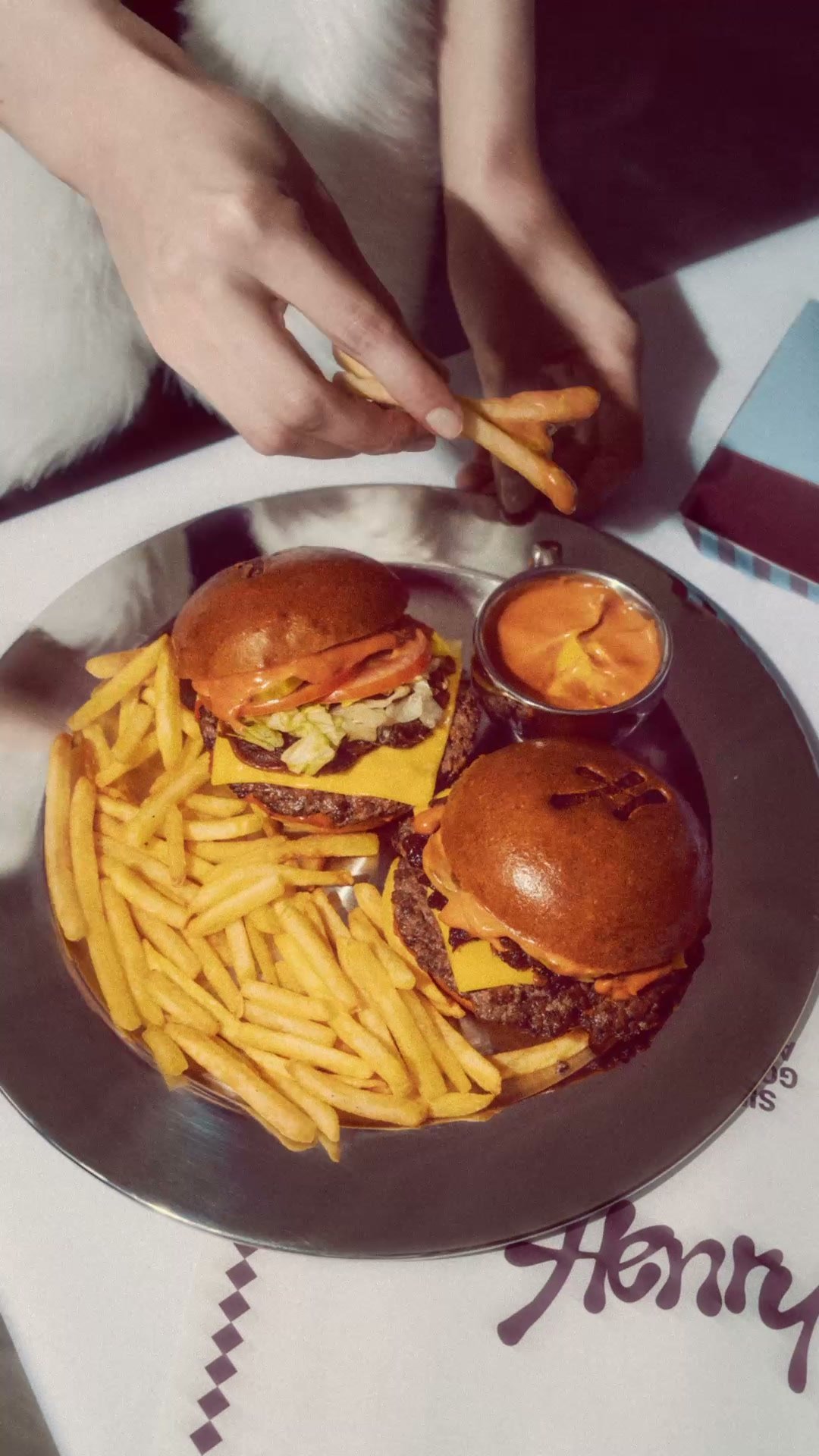 Hands taking fries from a plate of sliders and fries on a Henry tablecloth