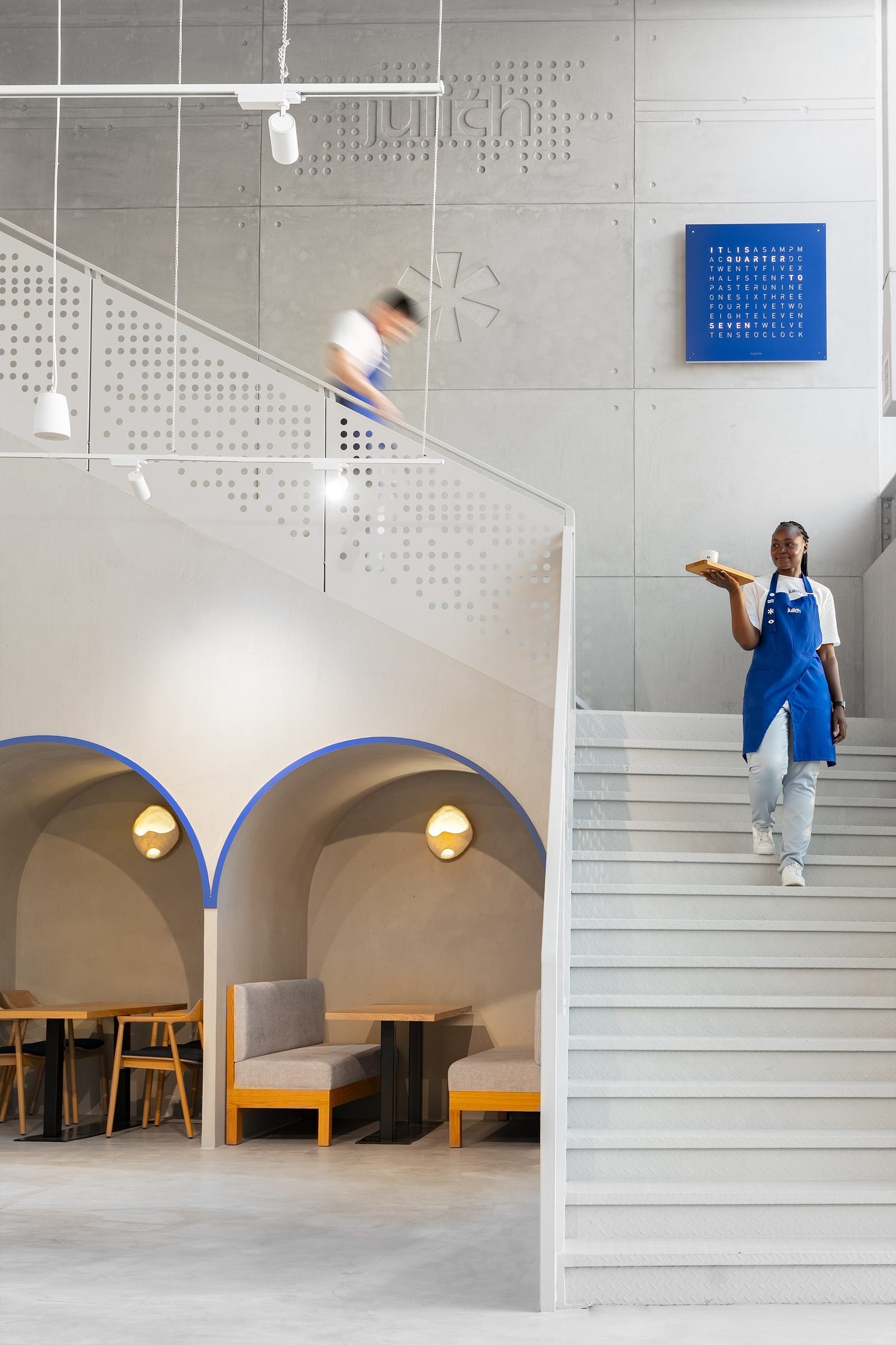 Woman in a blue apron on the stairs of a pale, arched cafe interior