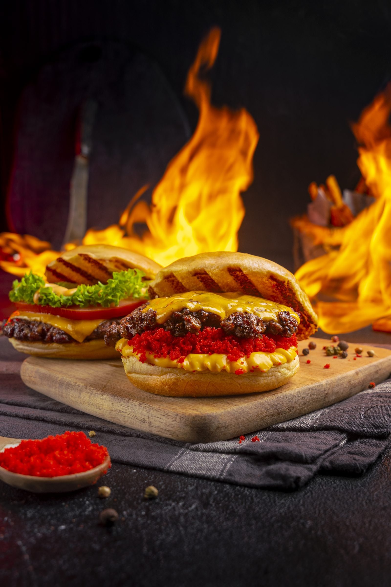 Two open burgers on a wooden board in front of open flames, with a spoon of chilli powder