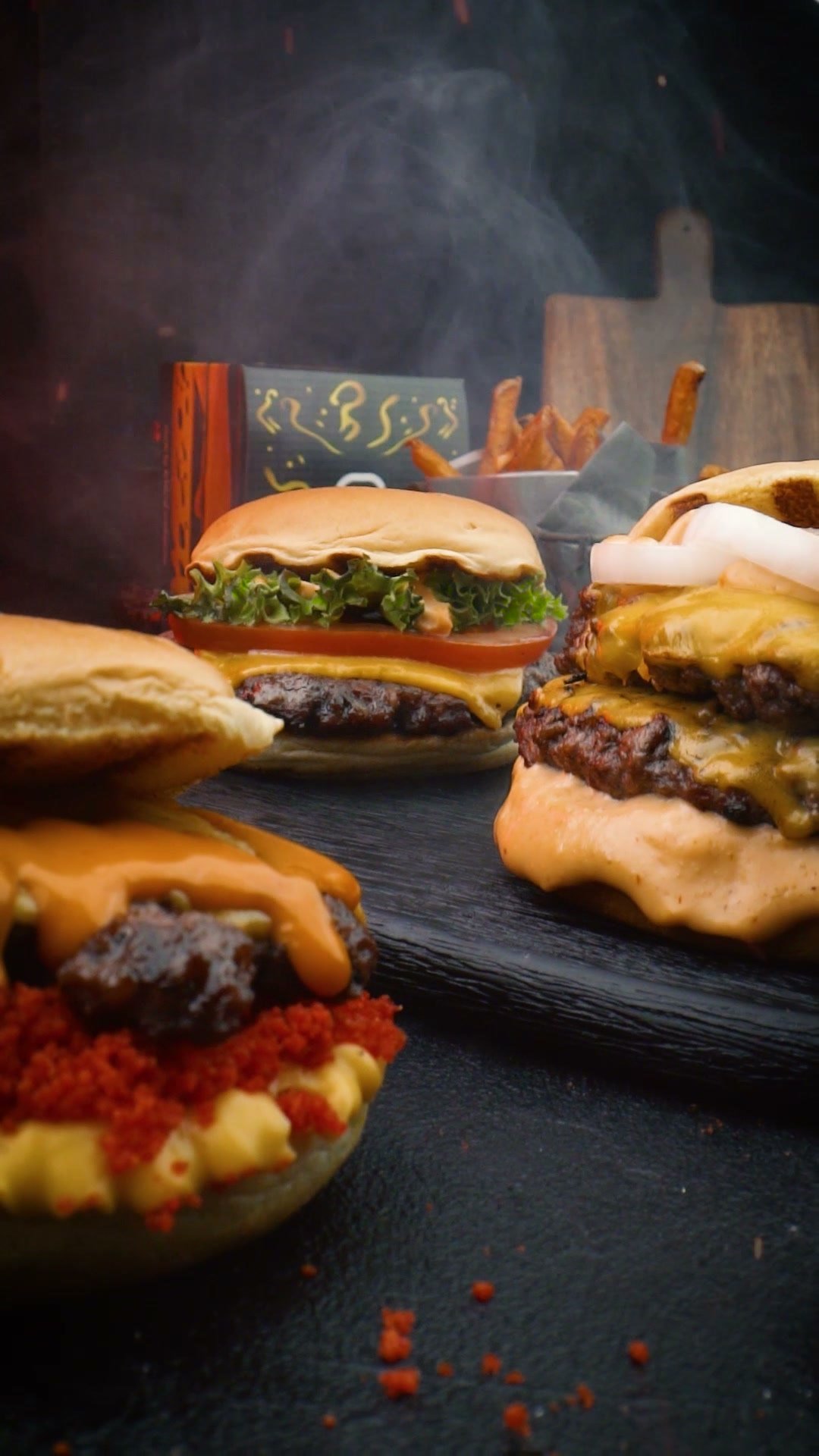 Three burgers with melted cheese and chilli on a dark counter, smoke behind them