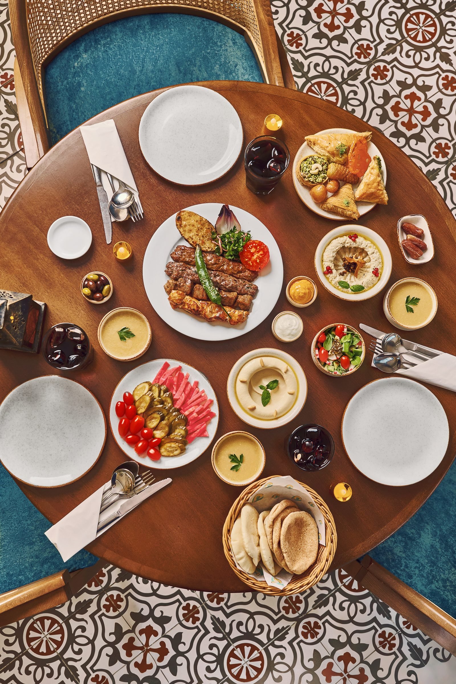 Overhead of a round table laid with mezze, grills and bread on a patterned tiled floor