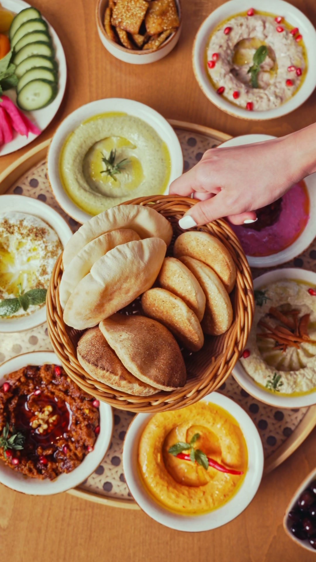 Overhead mezze spread of hummus, pickles, dips and a basket of breads, hands reaching in