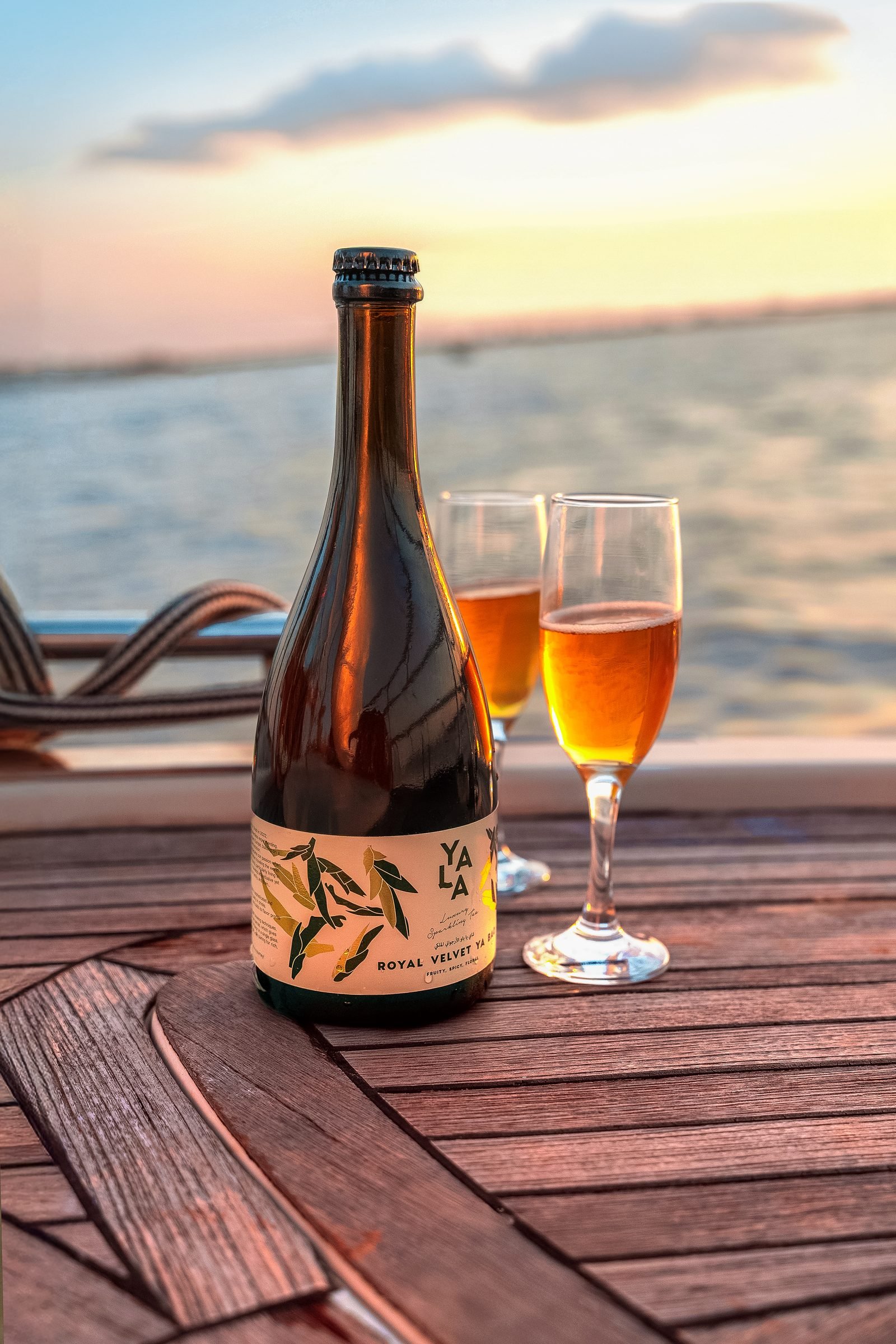 Bottle of kombucha and two filled glasses on a wooden deck at sunset by the water