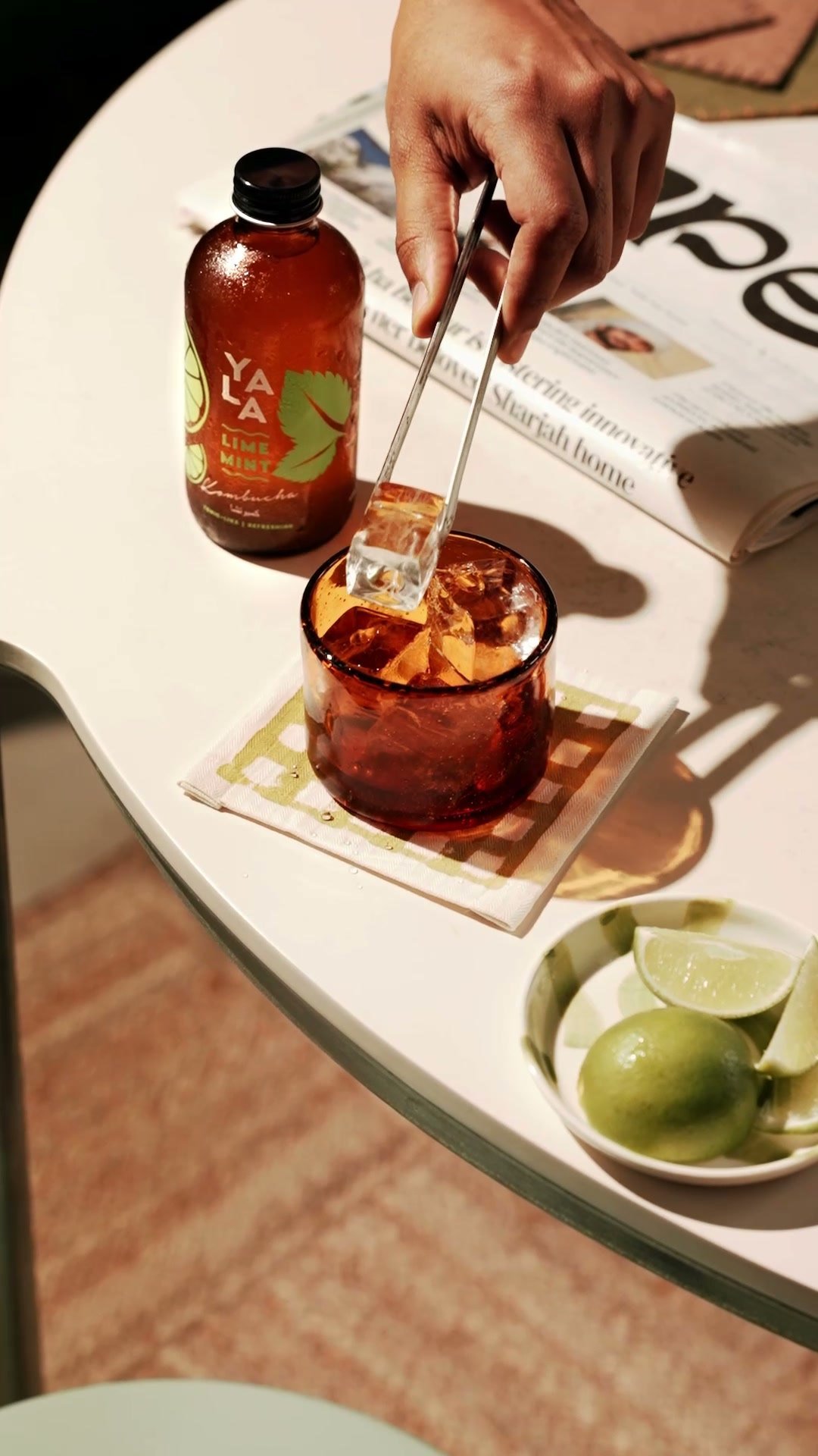 Hand stirring a glass of kombucha over ice on a marble table, limes and the bottle beside it