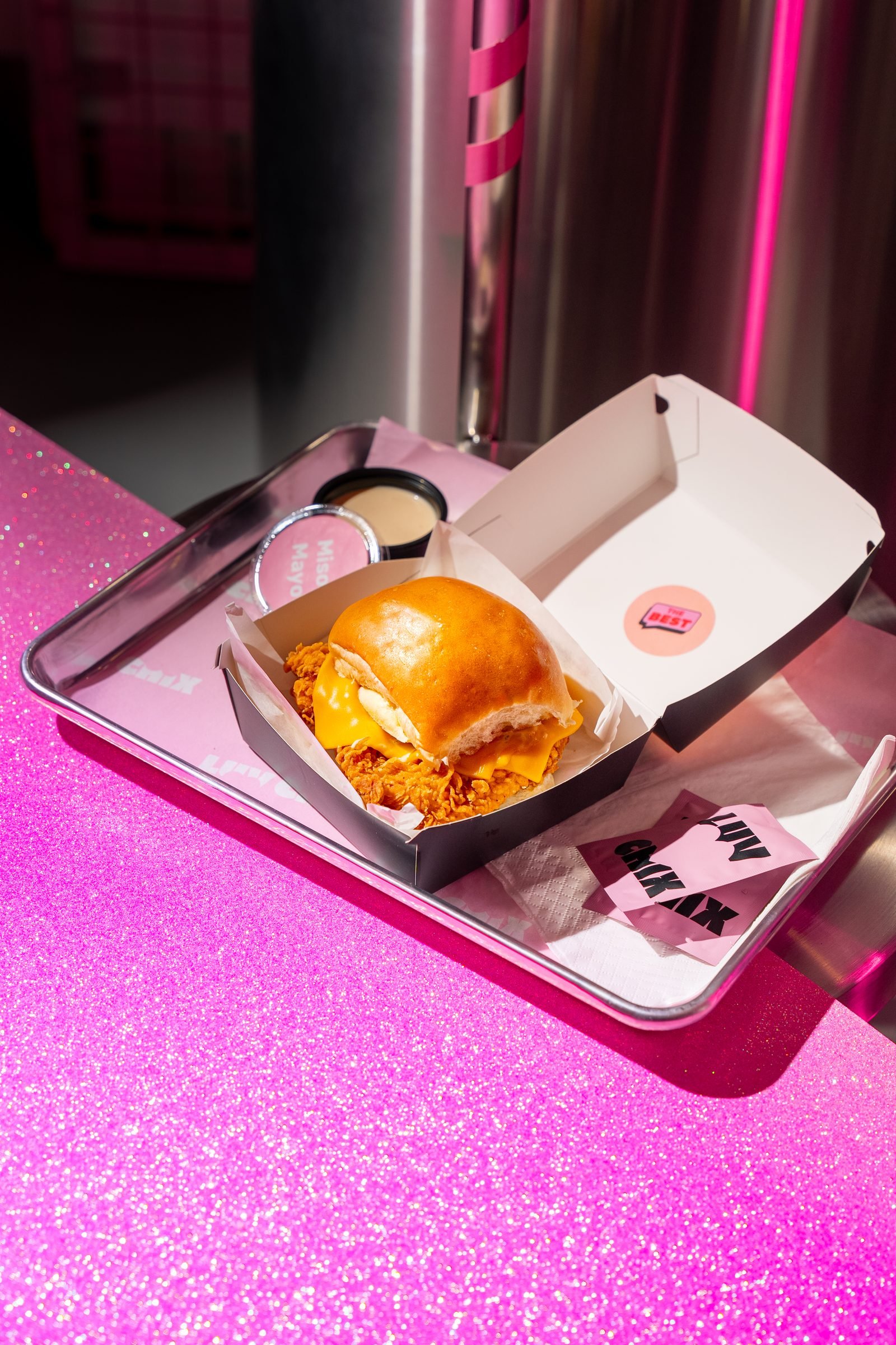 Chicken burger in an open box on a metal tray, lit magenta