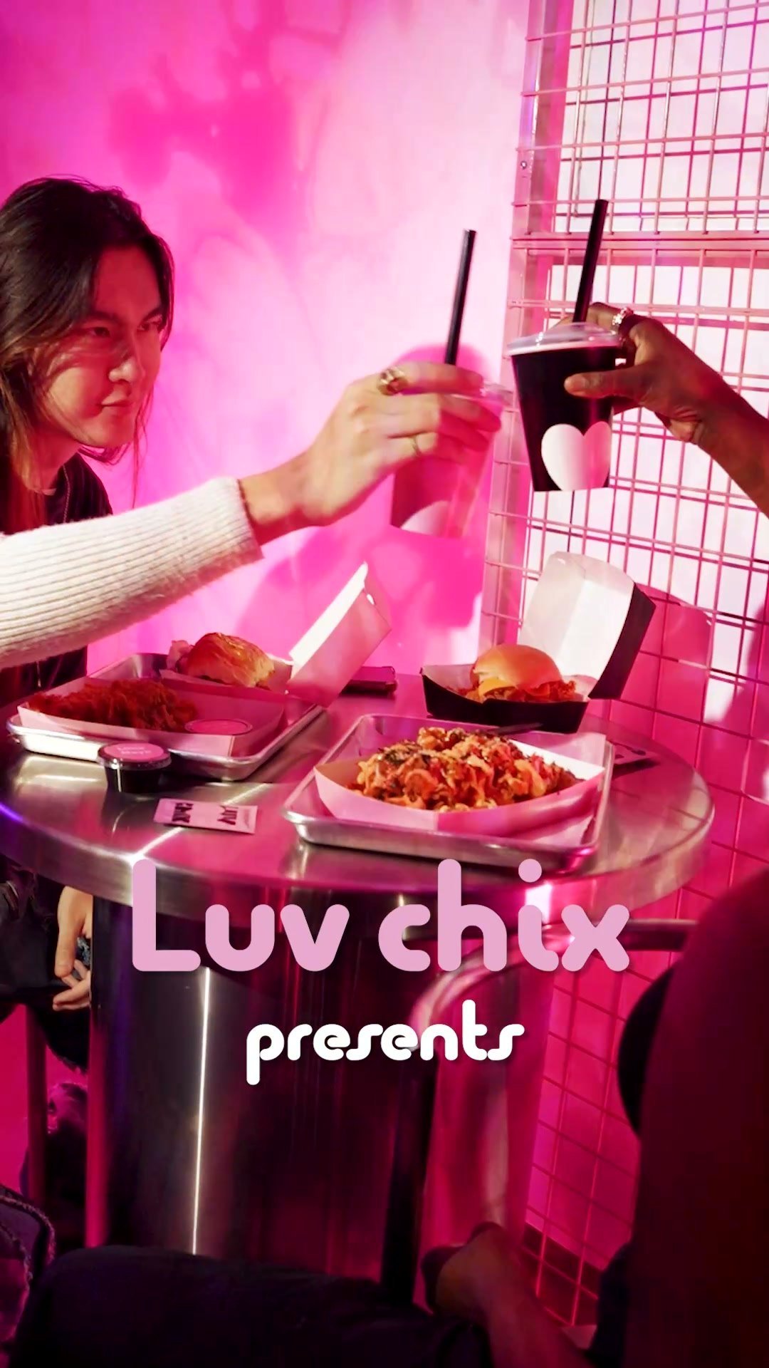 Two people toasting drinks over a spread of Luv Chix chicken and fries, in pink light