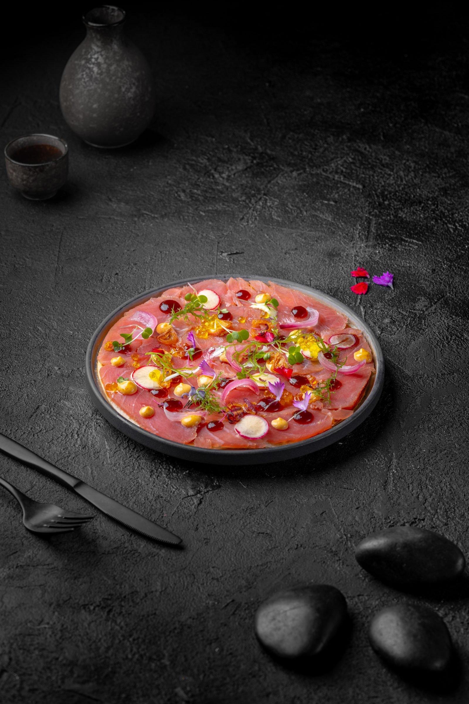 Round plate of thinly sliced fish with edible flowers and dots of sauce on dark stone