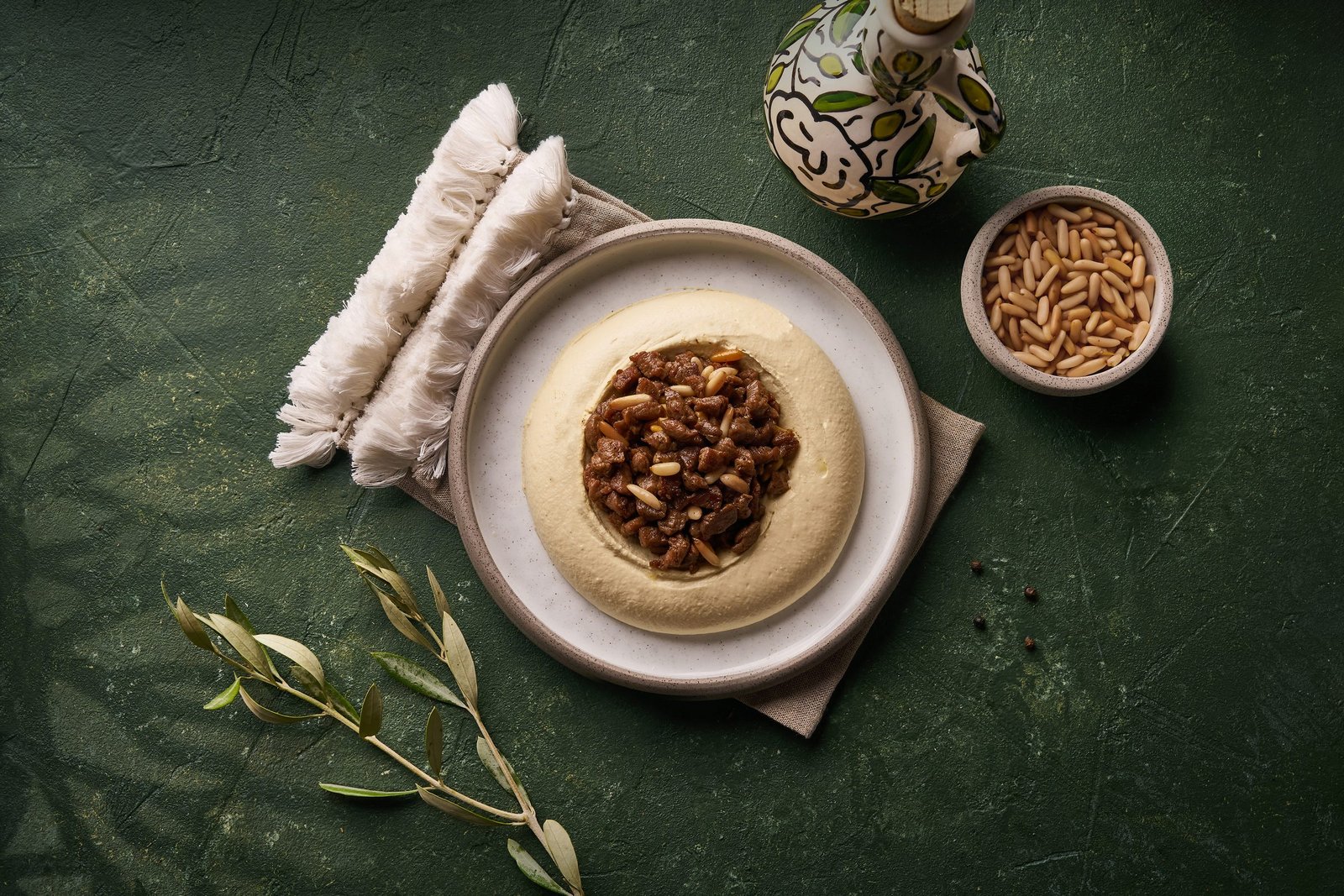 Hummus topped with minced meat and pine nuts, wrapped bread and an olive sprig on green