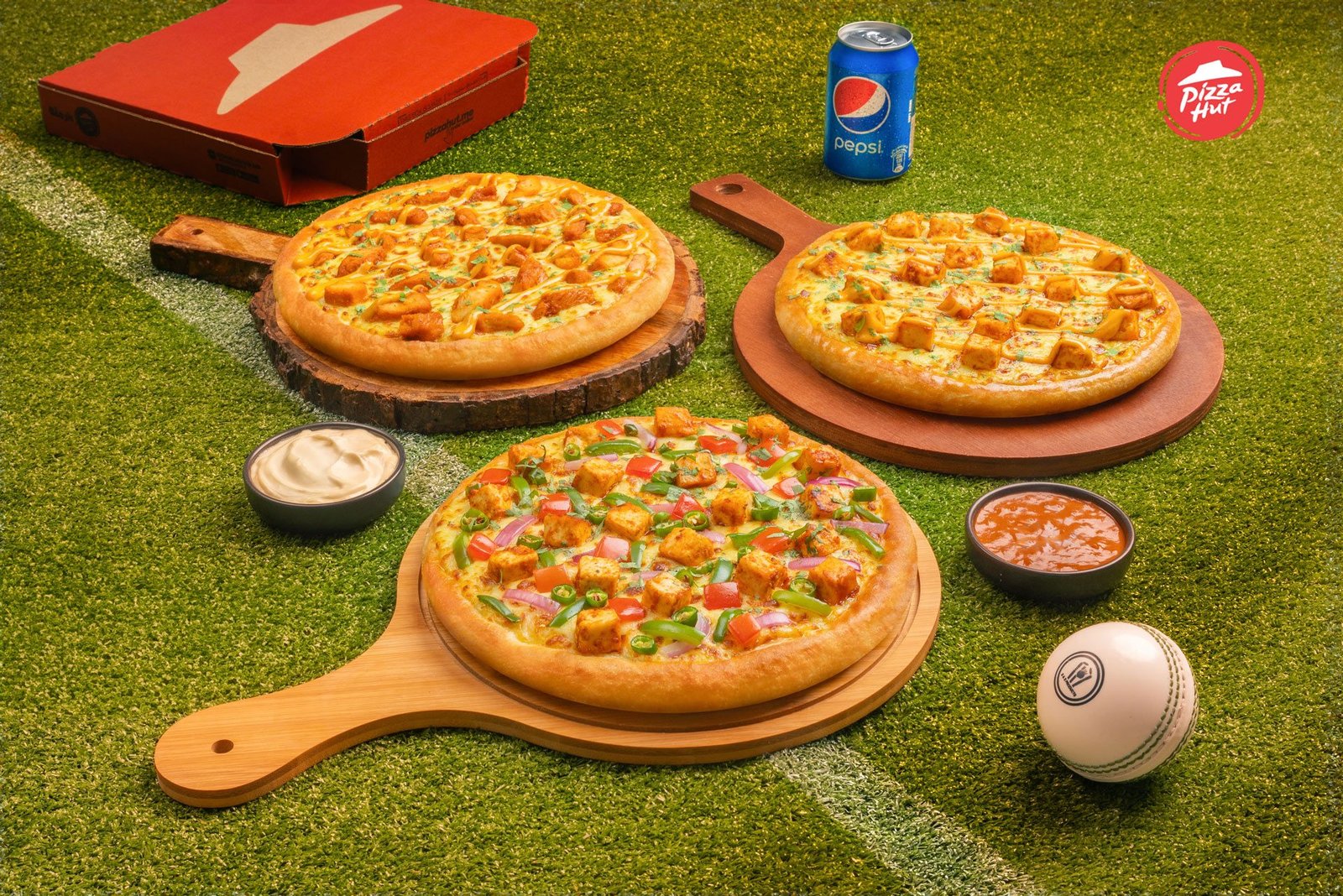 Three pizzas on wooden paddles laid out on a cricket pitch with a Pepsi can and a cricket ball