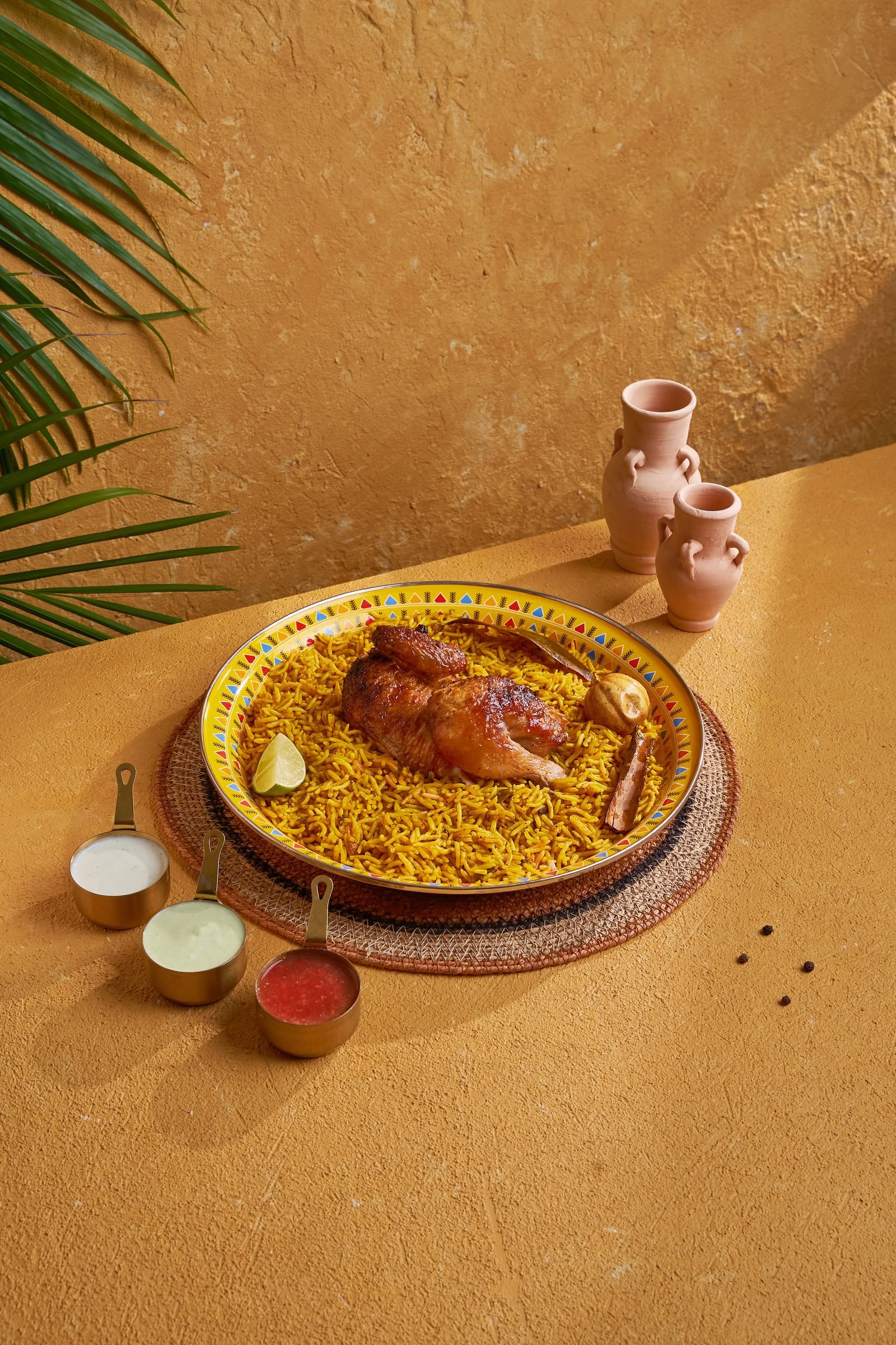 Roast chicken on saffron rice on a patterned platter, beside clay water jugs and palm shadow