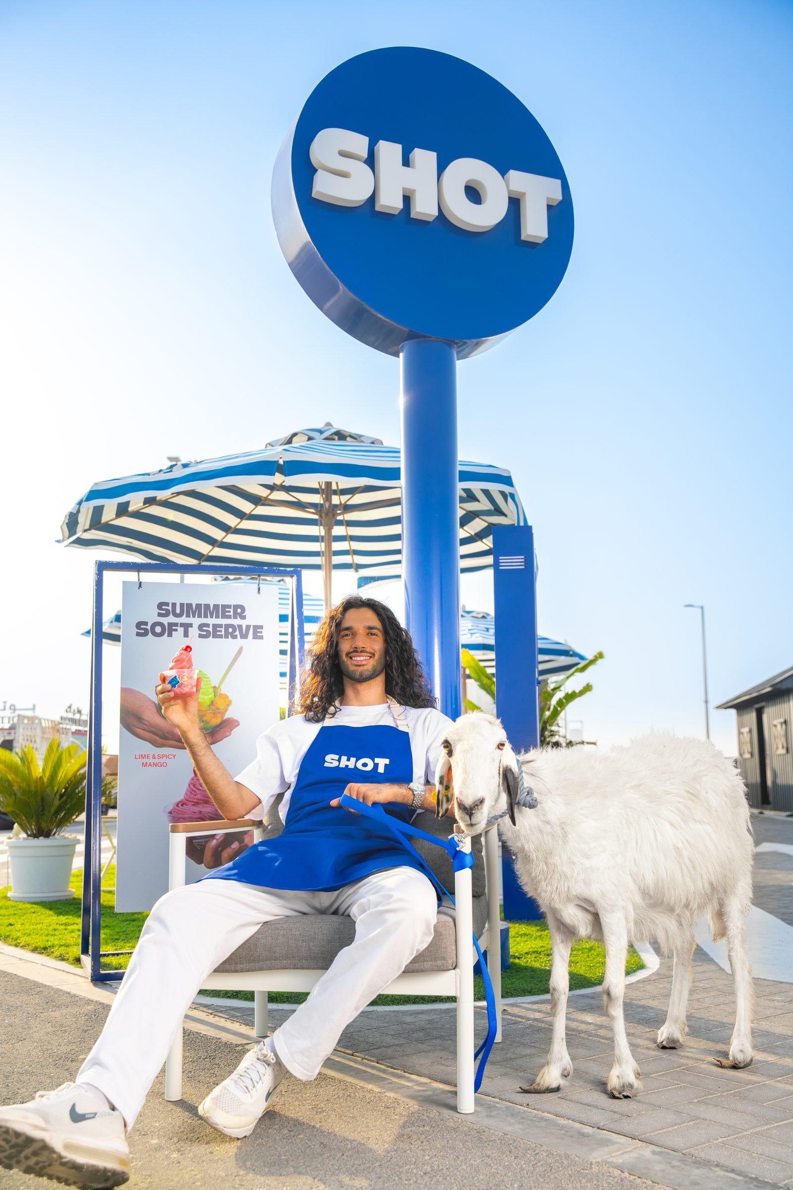 Woman seated under a Shot sign on a promenade, a goat standing beside her