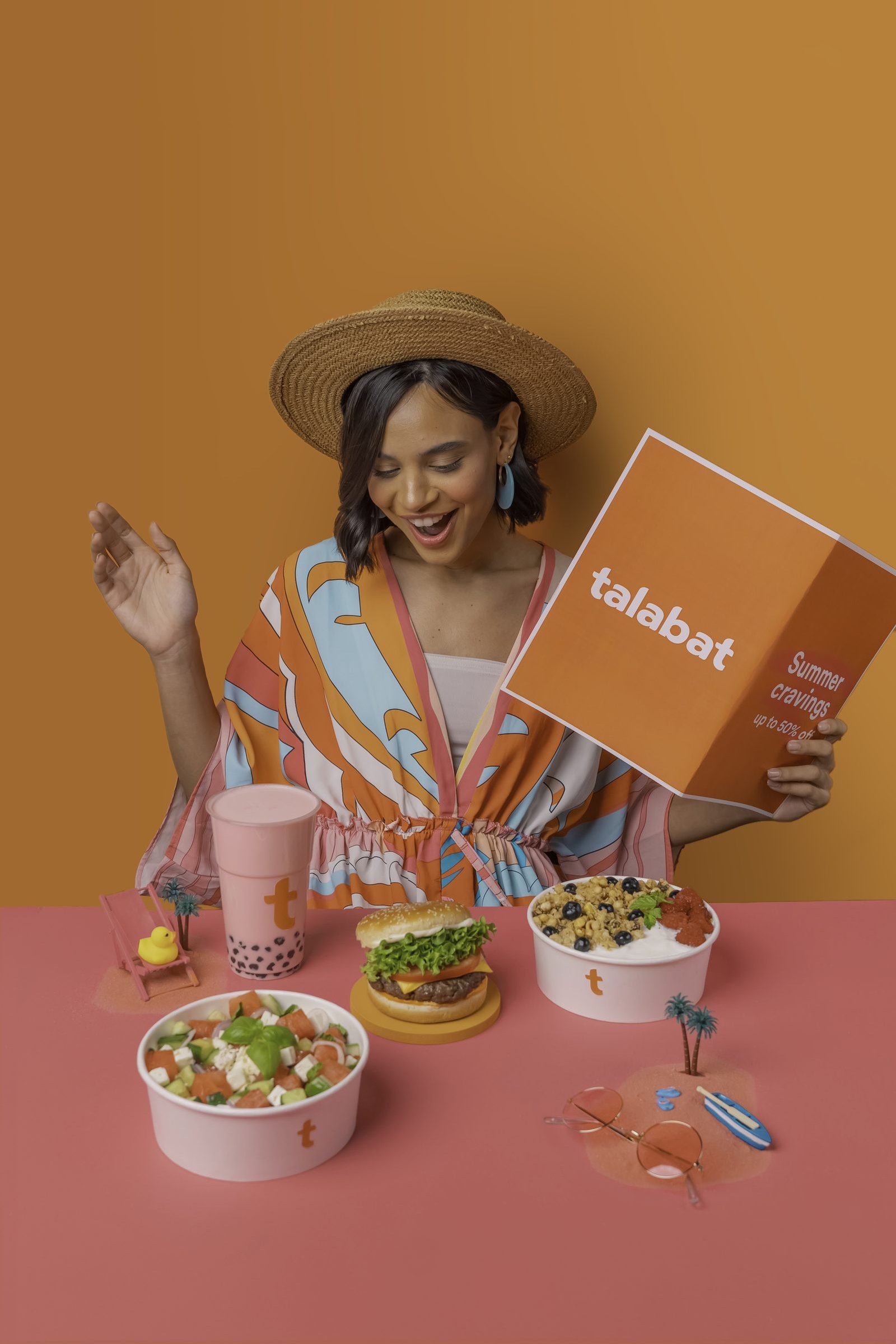 Smiling woman in a straw hat holding a Talabat box above bowls of salad and iced drinks