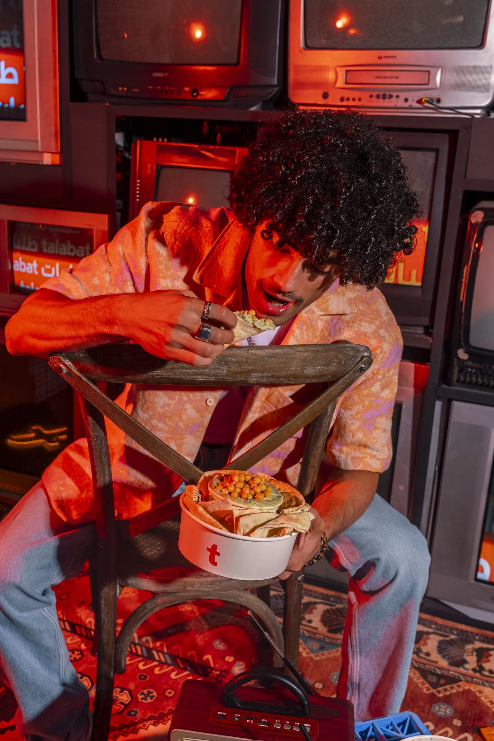 Man in an orange shirt eating from a takeaway bowl in front of a wall of glowing vintage televisions