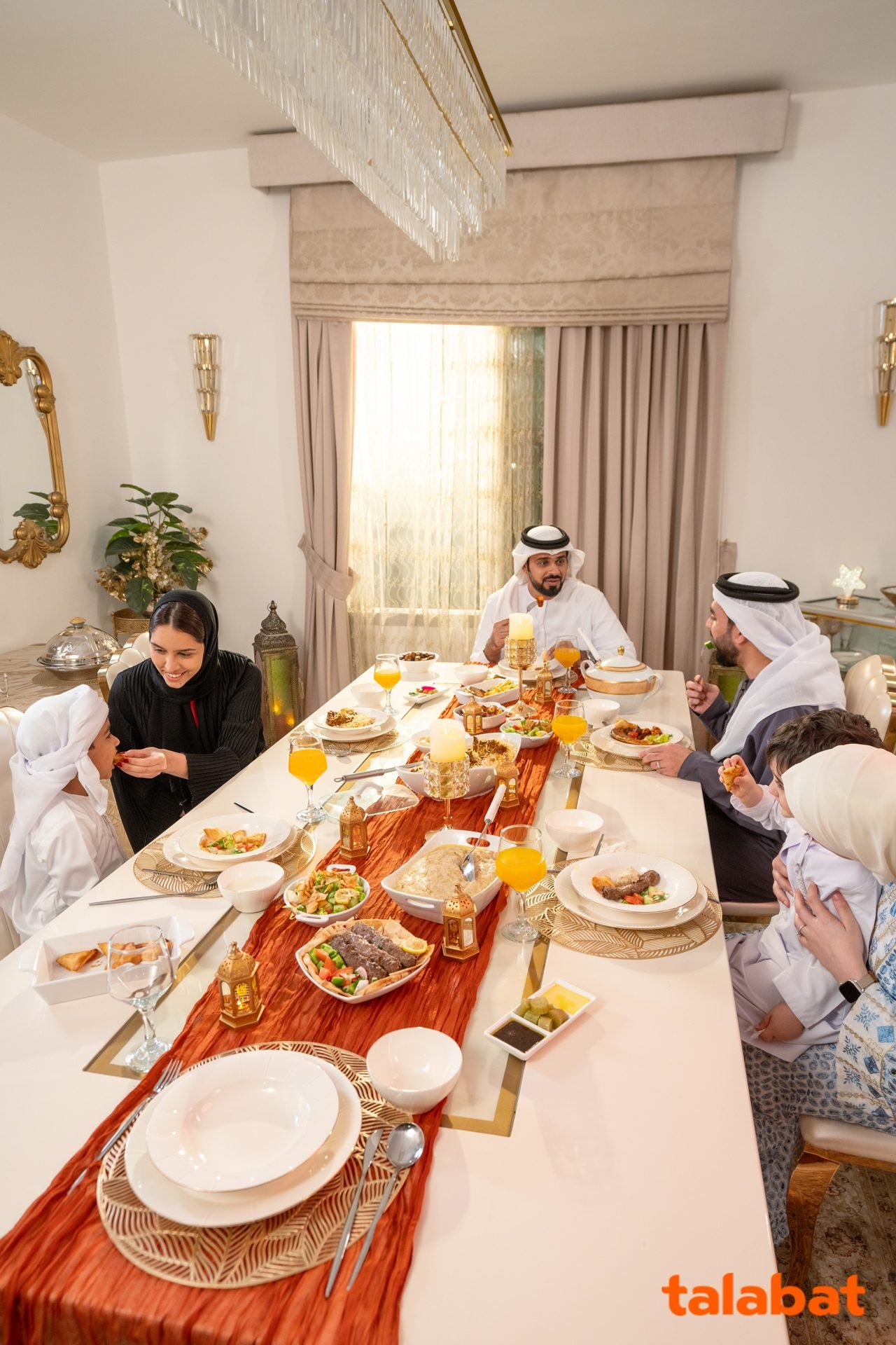 Long family table laid with Emirati dishes and juice, guests in kanduras and abayas seated along it