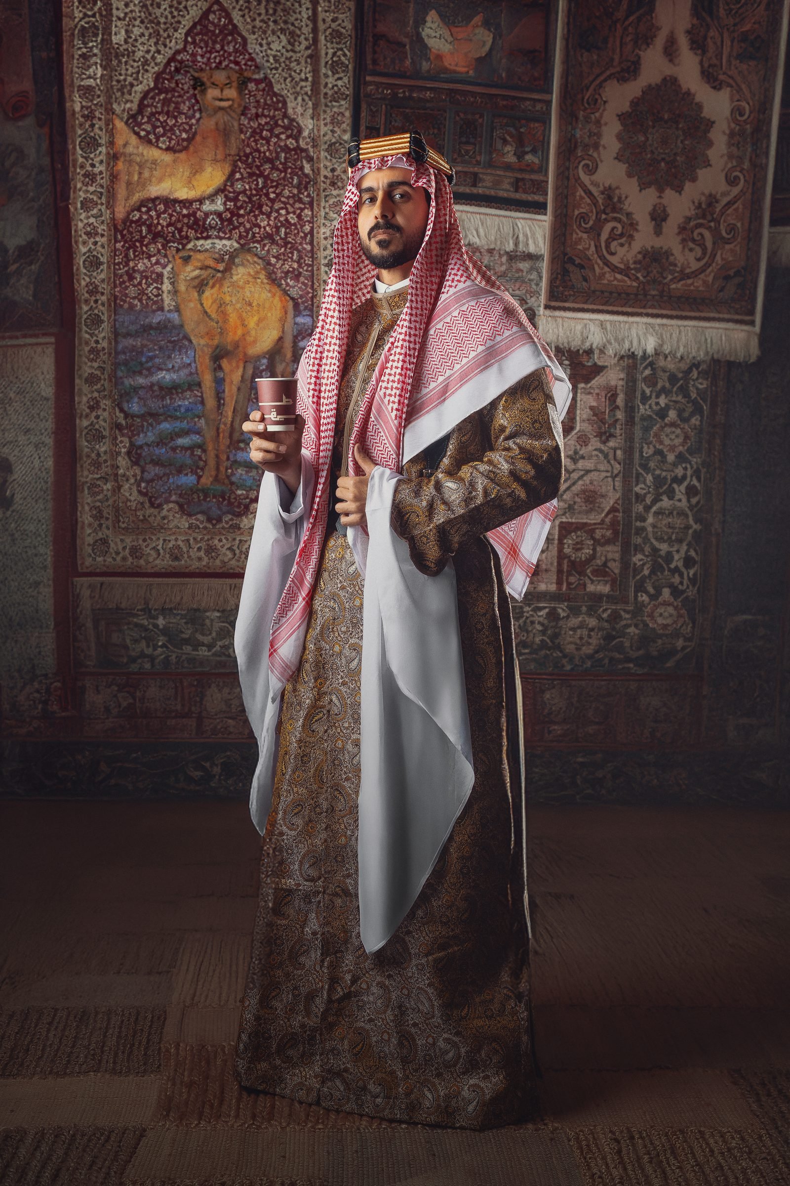 Man in a bisht and ghutra holding a takeaway cup in front of a carpeted majlis wall