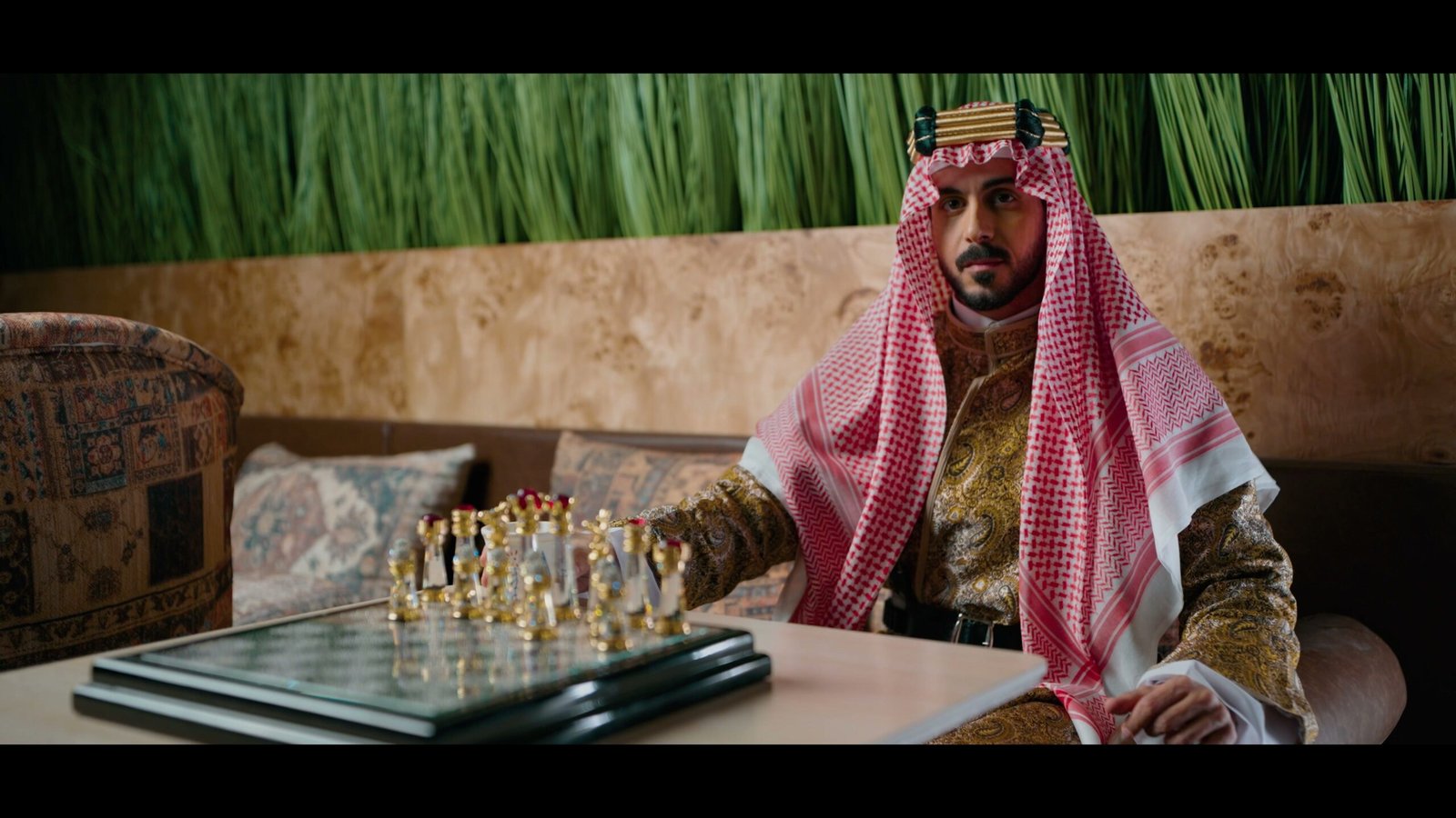 Man in Saudi dress seated at a low table with a chess set, against a wall of green grass