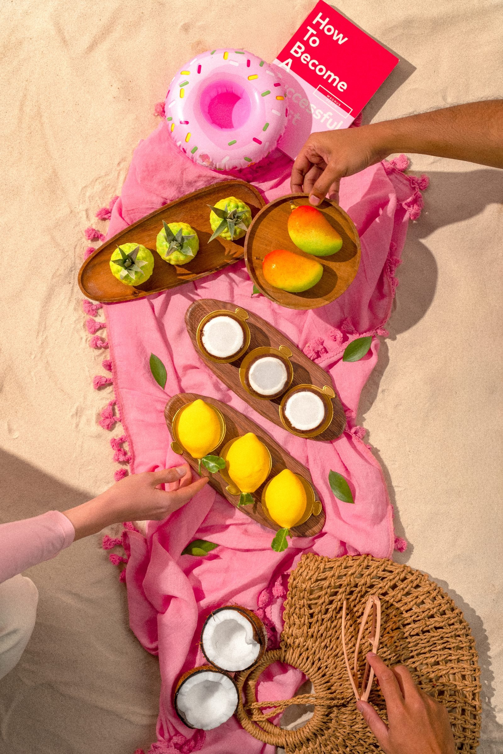Overhead of fruit tarts and lemons on a pink beach towel, hands setting down a straw bag