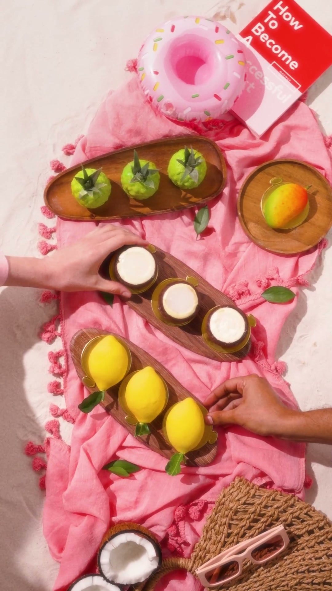 Overhead of fruit tarts, lemons and a coconut on a pink beach towel, hands arranging them
