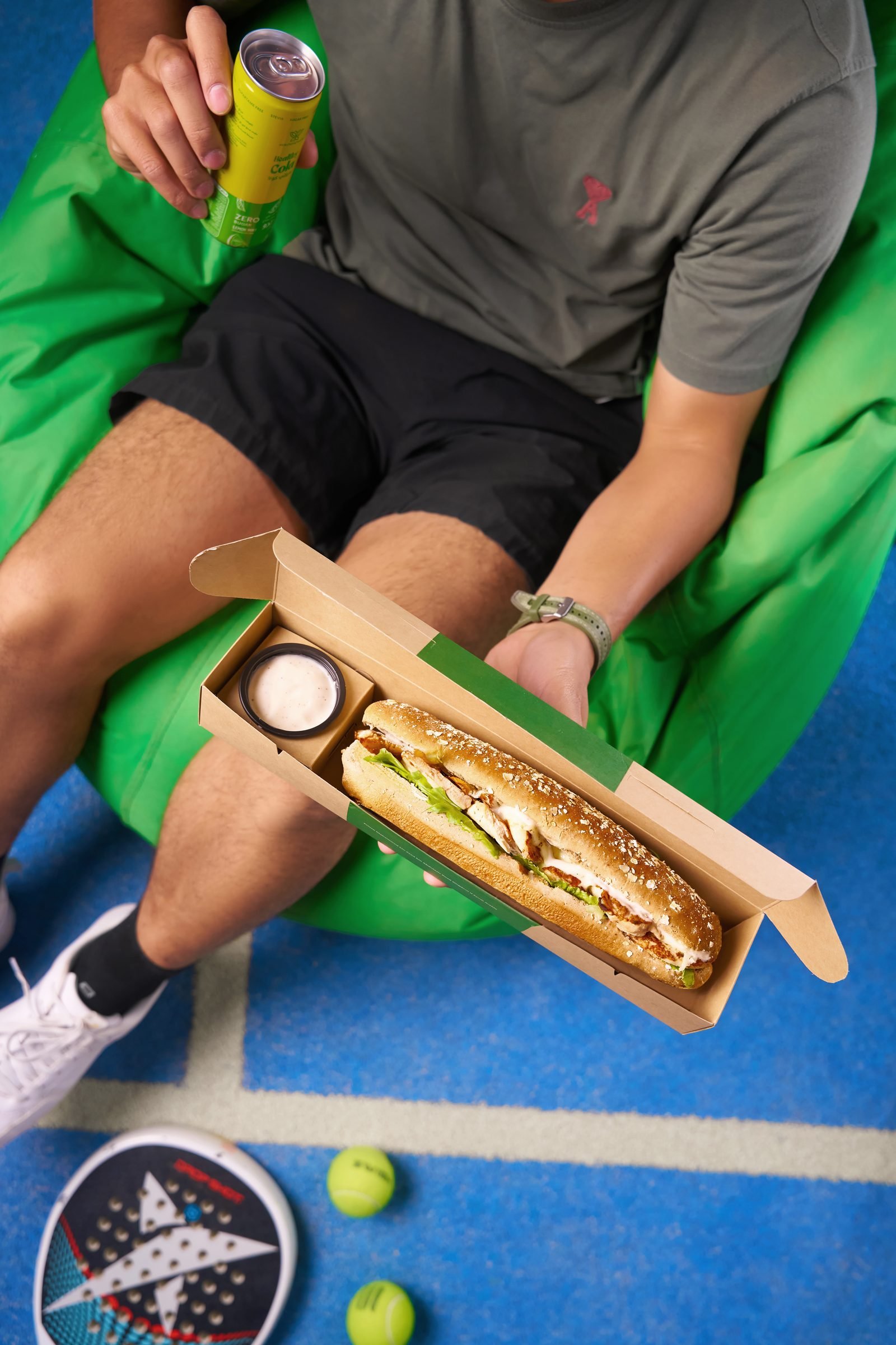 Man on a green beanbag on an outdoor court opening a Well Health box with a baguette inside