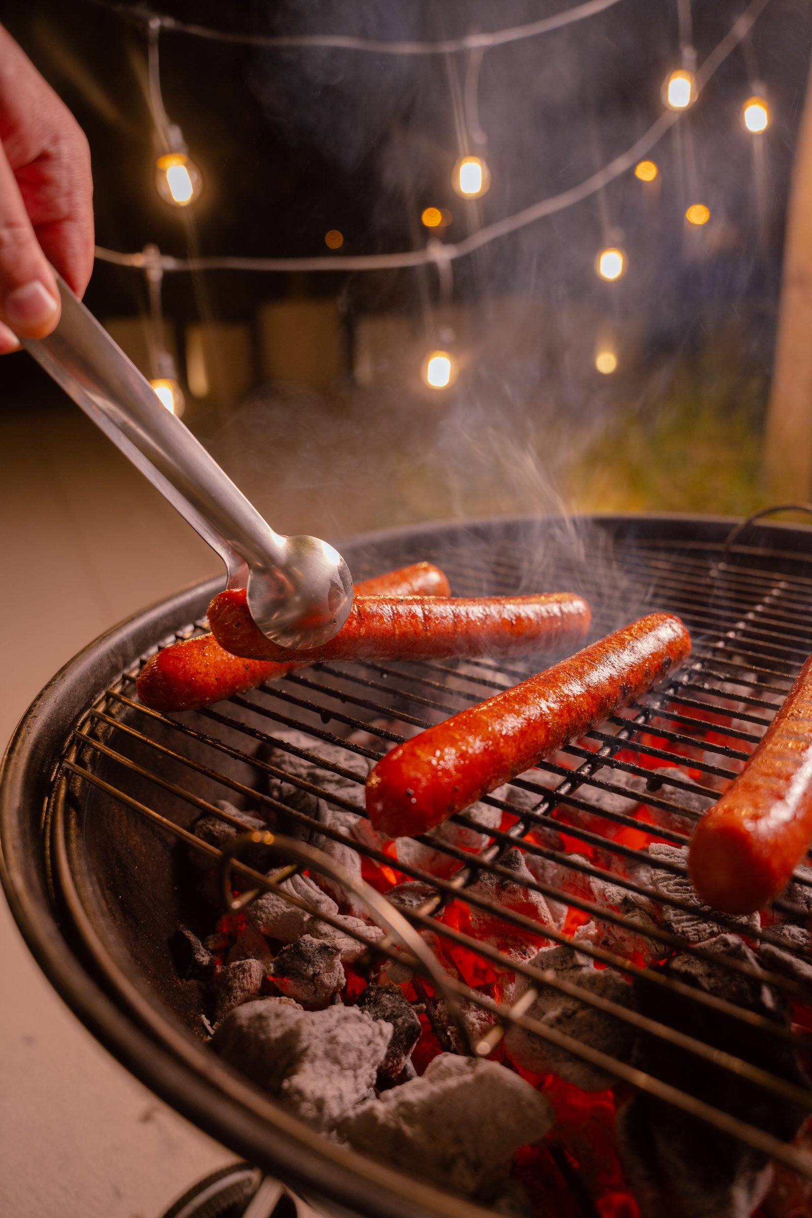Tongs turning sausages on a charcoal grill under string lights
